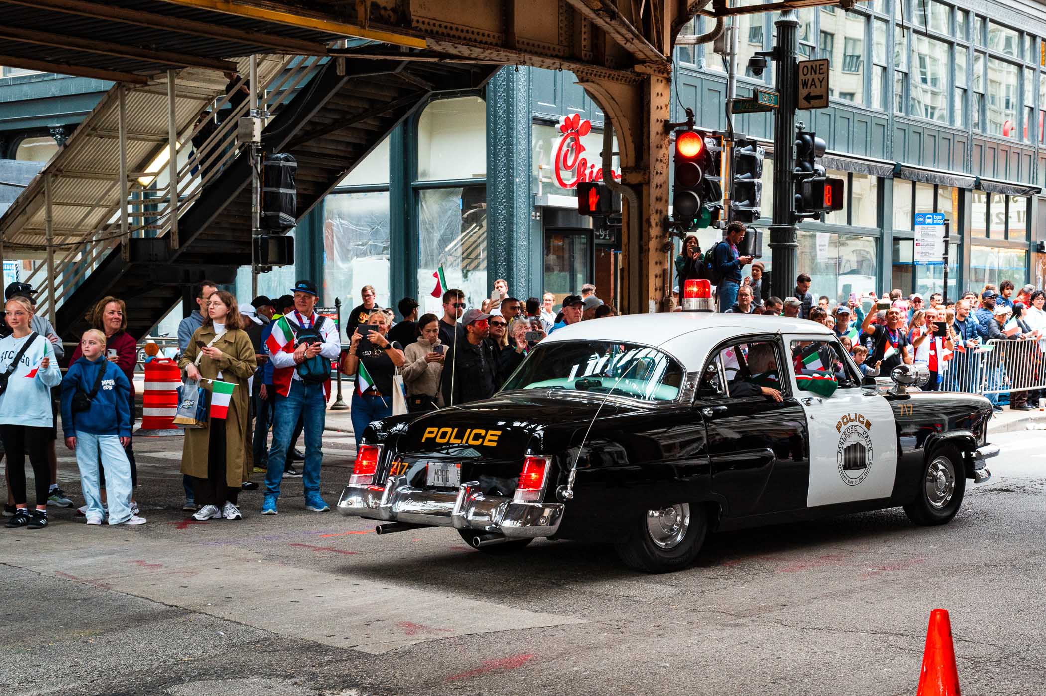 A vintage police car with its lights on drives past a crowd celebrating Columbus Day in Chicago, featuring people holding Italian flags and enjoying the event.