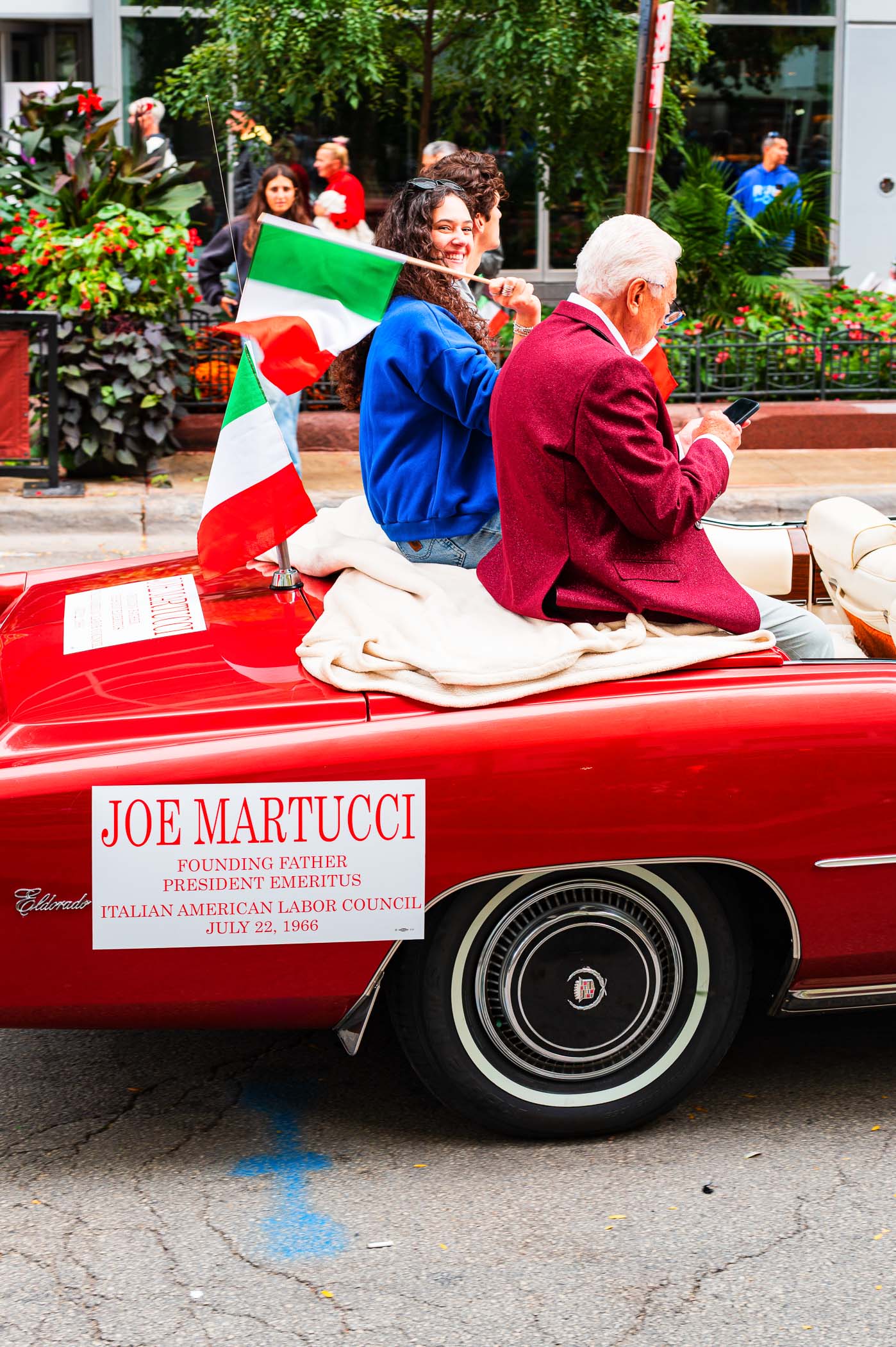 A young woman smiles and waves while holding an Italian flag, sitting next to an elderly man in a red blazer, both in a convertible car with a sign that reads 'Joe Martucci, Founding Father, President Emeritus, Italian American Labor Council'.