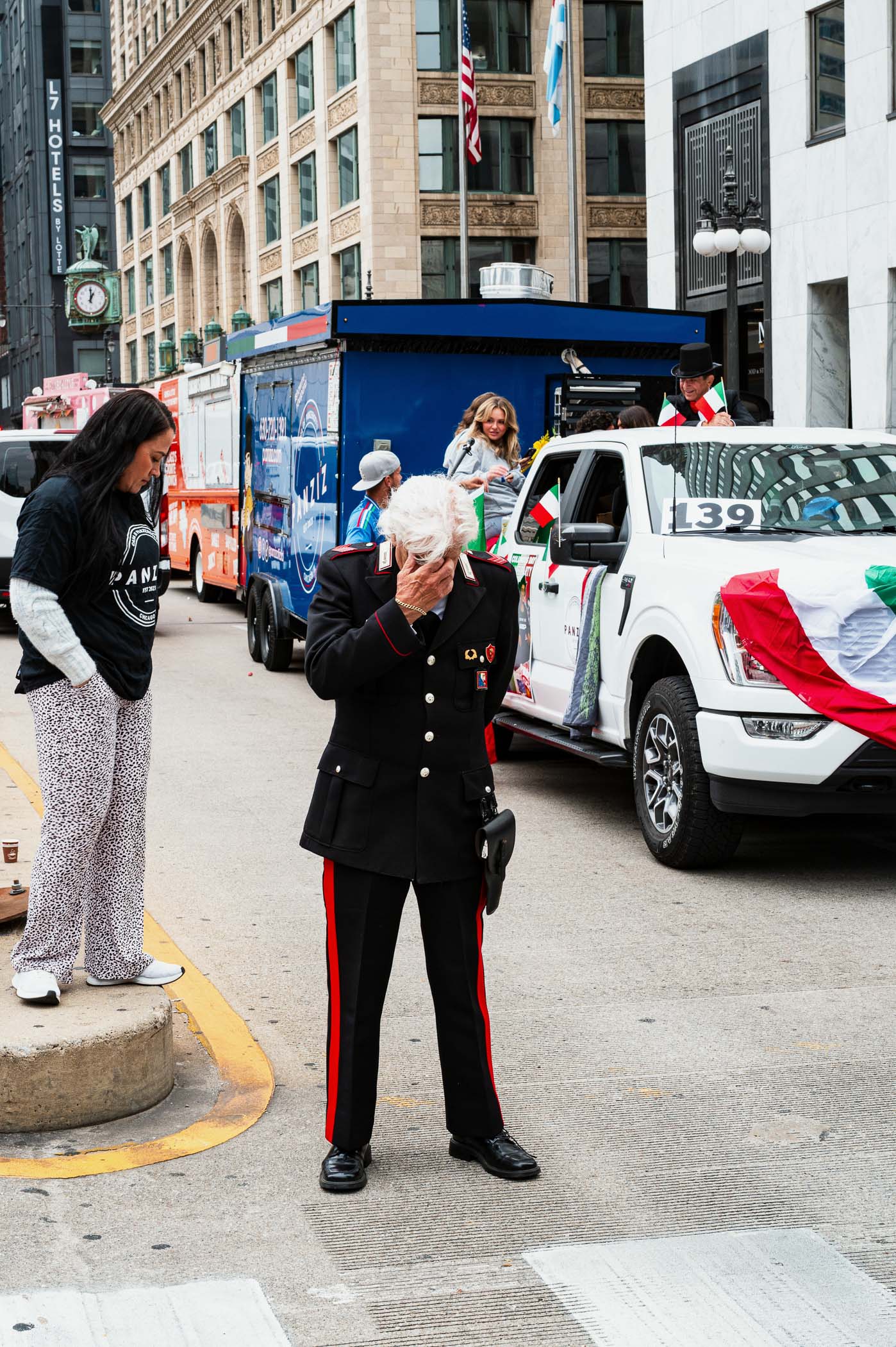 A parade scene on Columbus Day in Chicago, featuring a man in a military-style uniform with white hair, standing with his face covered, expressing emotion. Nearby, a woman in casual attire watches, alongside colorful decorated vehicles in the background.