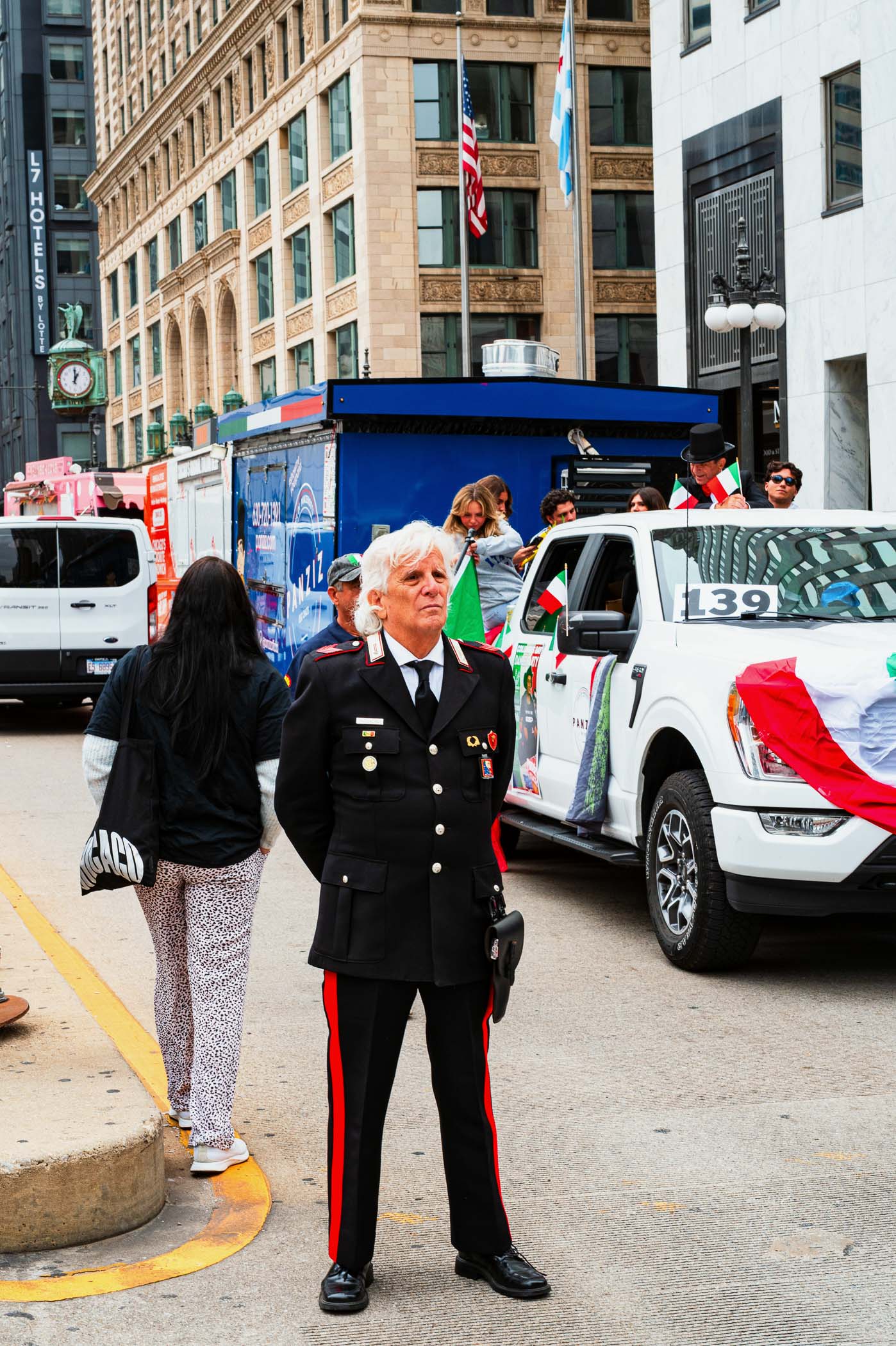 A police officer in a black uniform with red stripes stands in front of decorated vehicles at a Columbus Day parade in Chicago, surrounded by people celebrating.