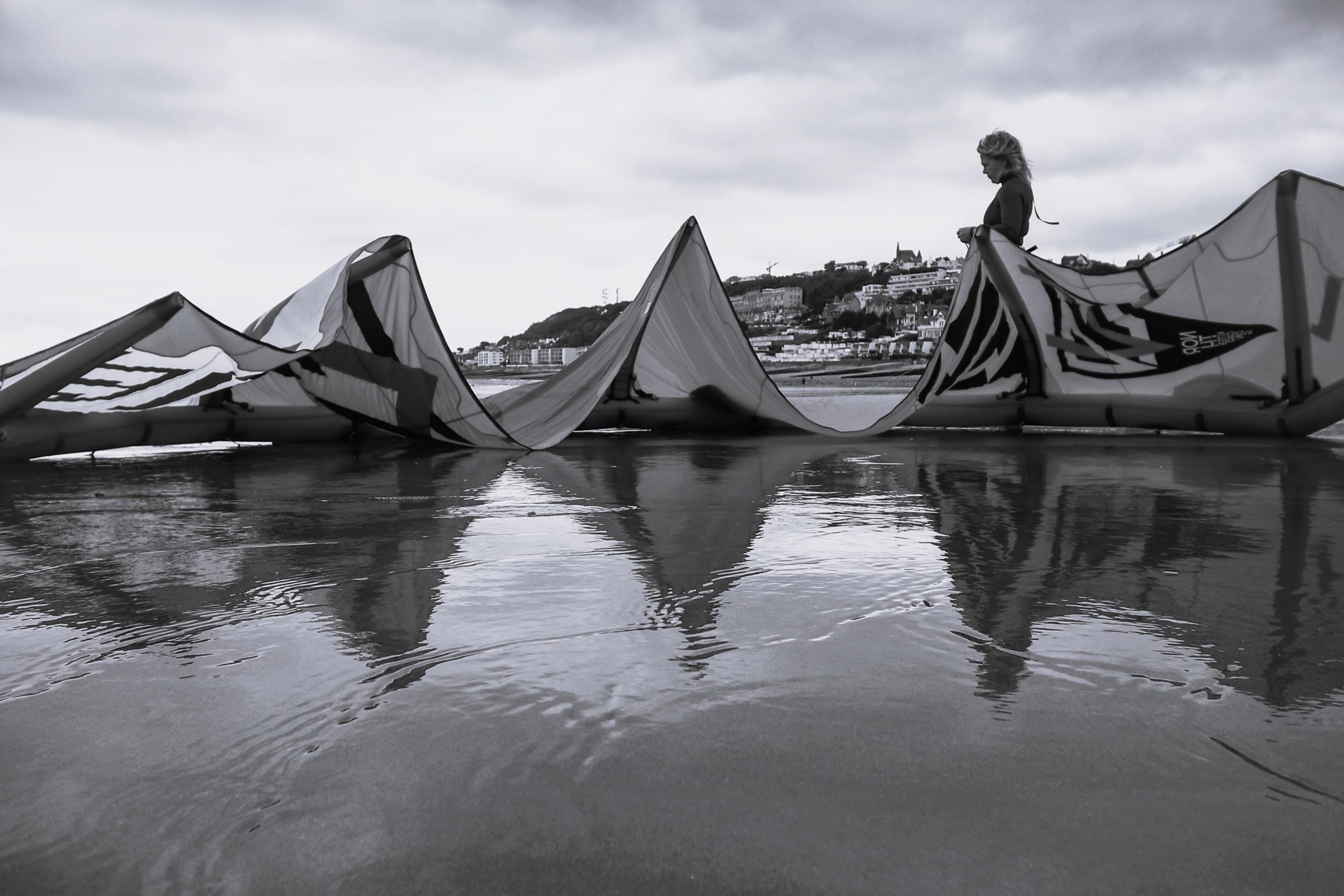 A black and white image of a kitesurfing kite draped across a wet beach, with reflections visible in the water. A person stands in the background, overlooking the beach and townscape.