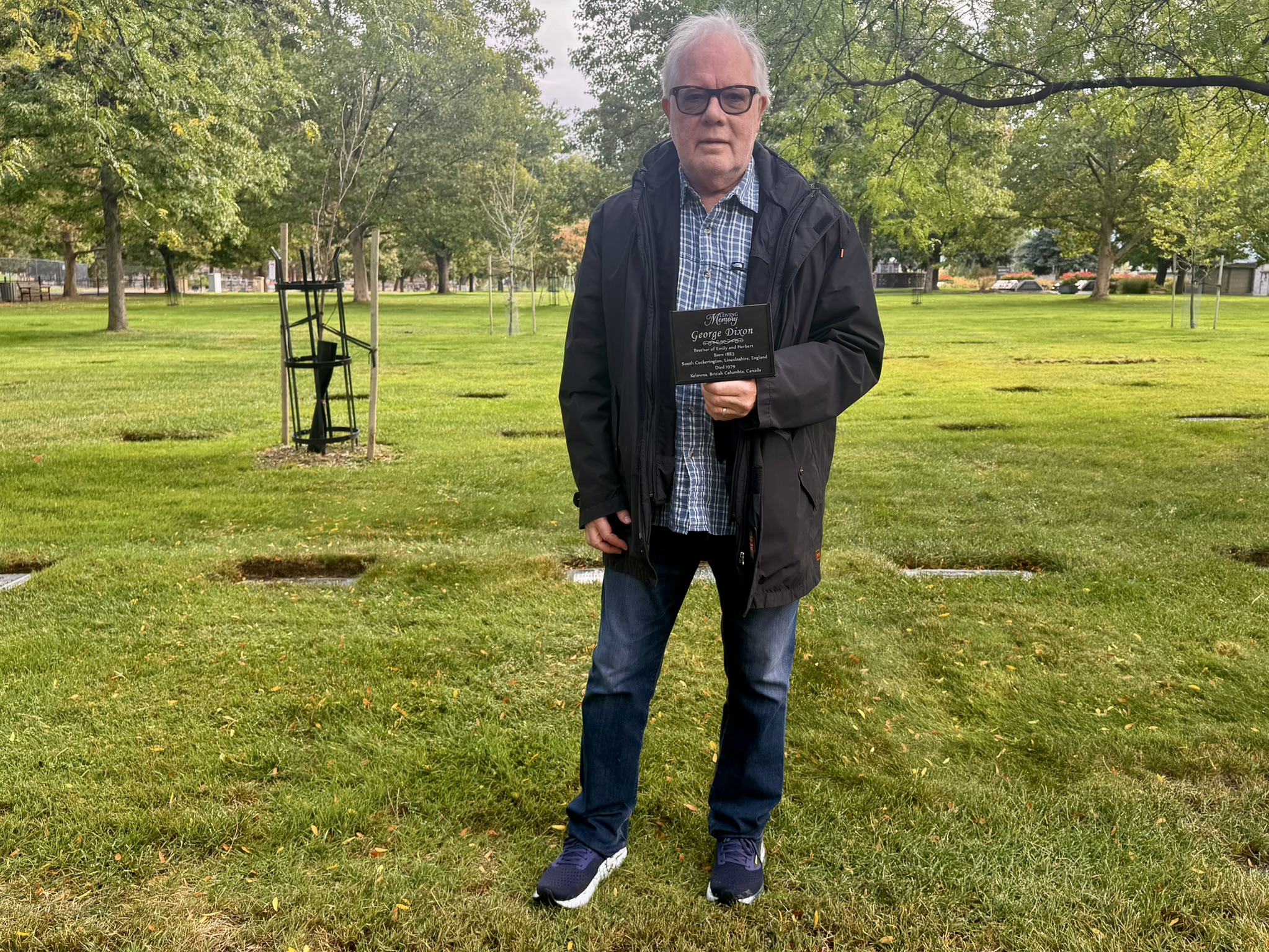 Man standing in a cemetery holding a gravestone plaque for George Dixon, with grassy surroundings and trees in the background.