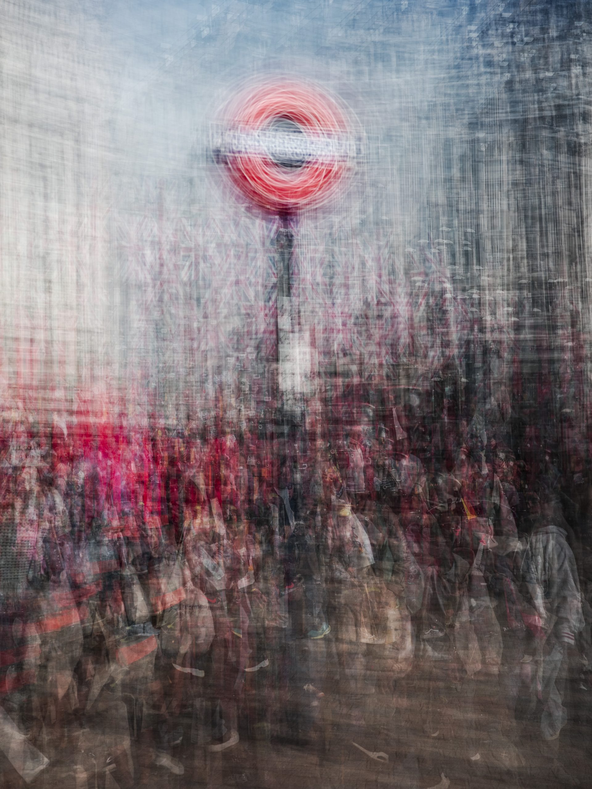 A blurred image of a busy urban scene featuring a prominent London Underground sign, amidst a crowd of people and colorful surroundings.