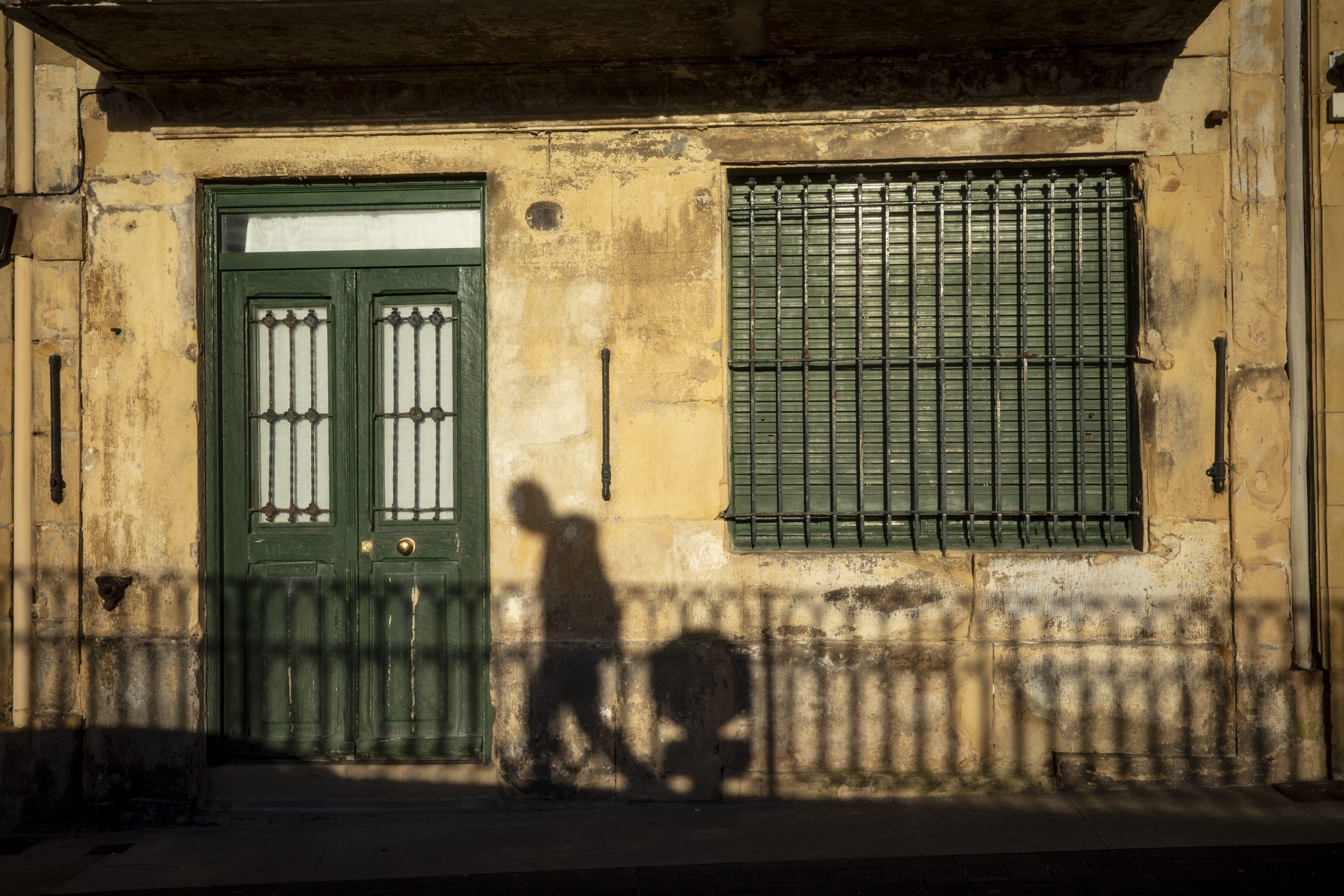 A shadow of a person walking with a stroller is cast against a textured yellow wall featuring a green door and barred window.