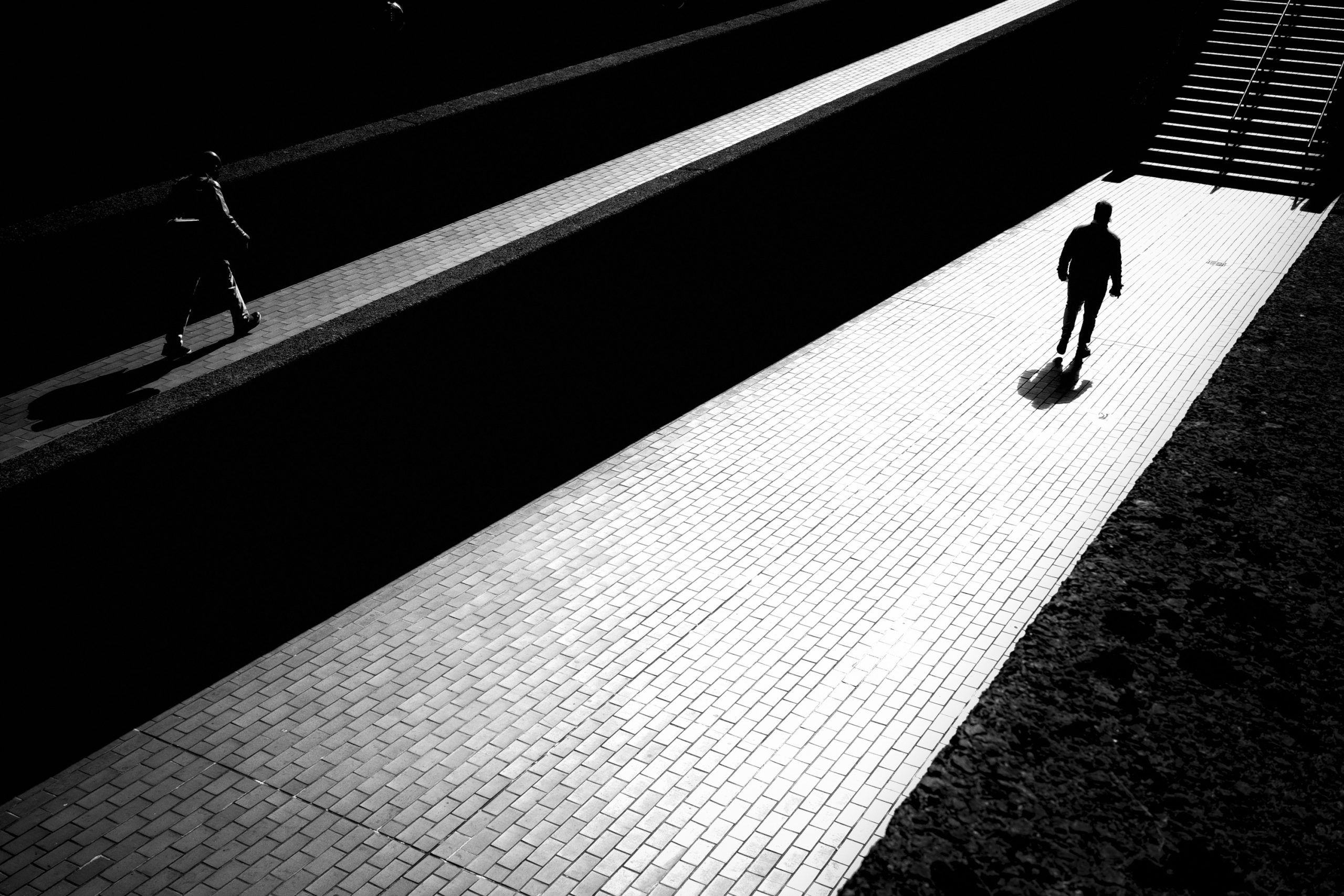 A black and white photograph capturing a lone figure walking along a bright pathway, contrasting sharply against dark surroundings, showcasing the interplay of light and shadow.