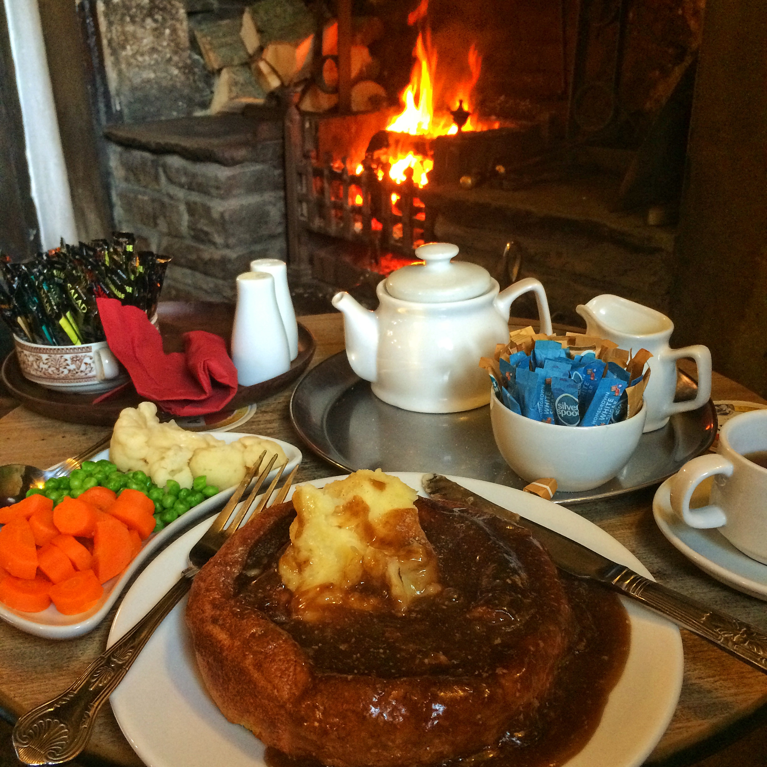 A hearty plate of Yorkshire roast beef served with Yorkshire pudding, vegetables including carrots, peas, and cauliflower, with a warm fireplace in the background.