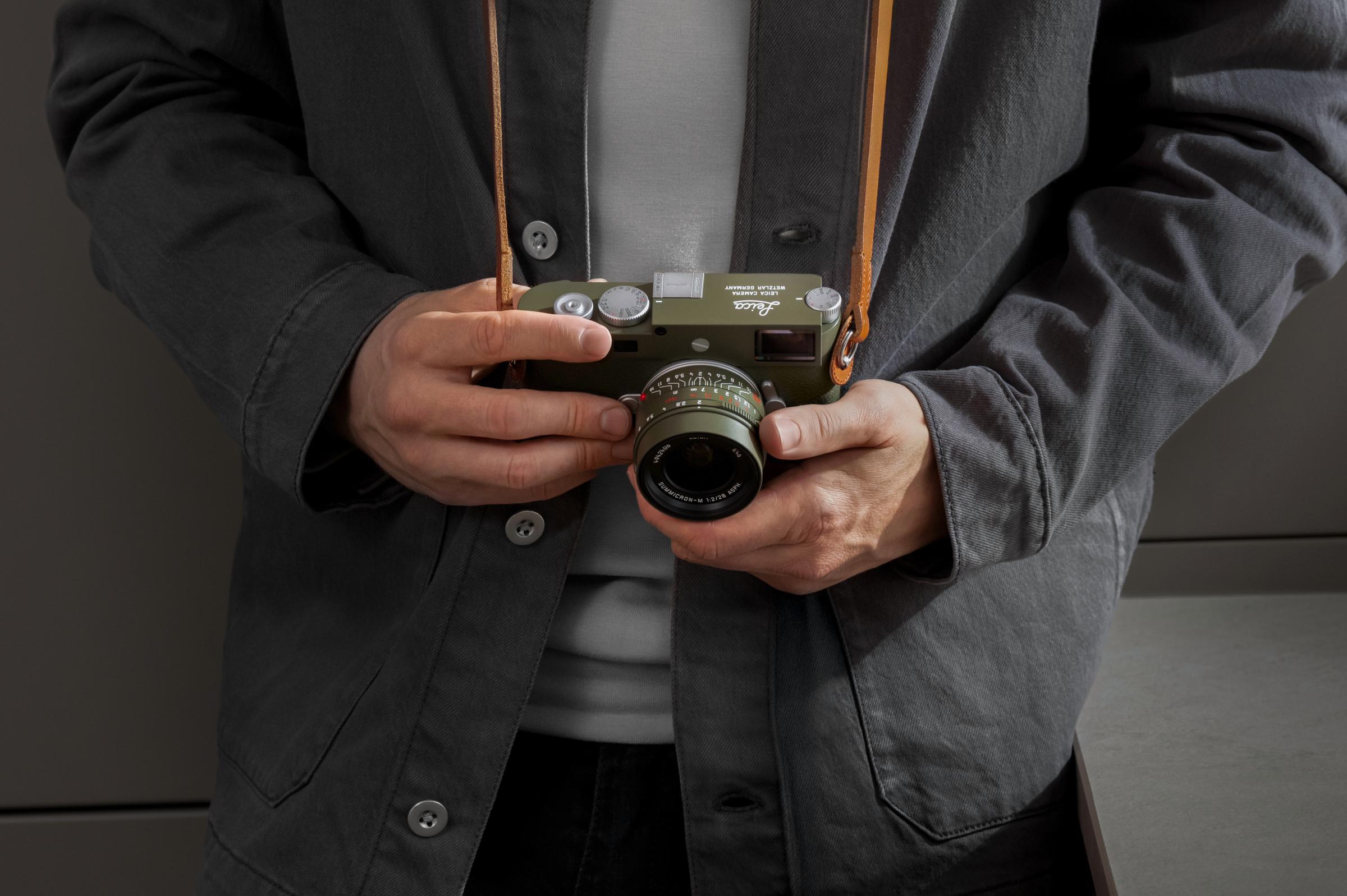 A person holding a Leica camera with a matte olive green finish, wearing a dark jacket.