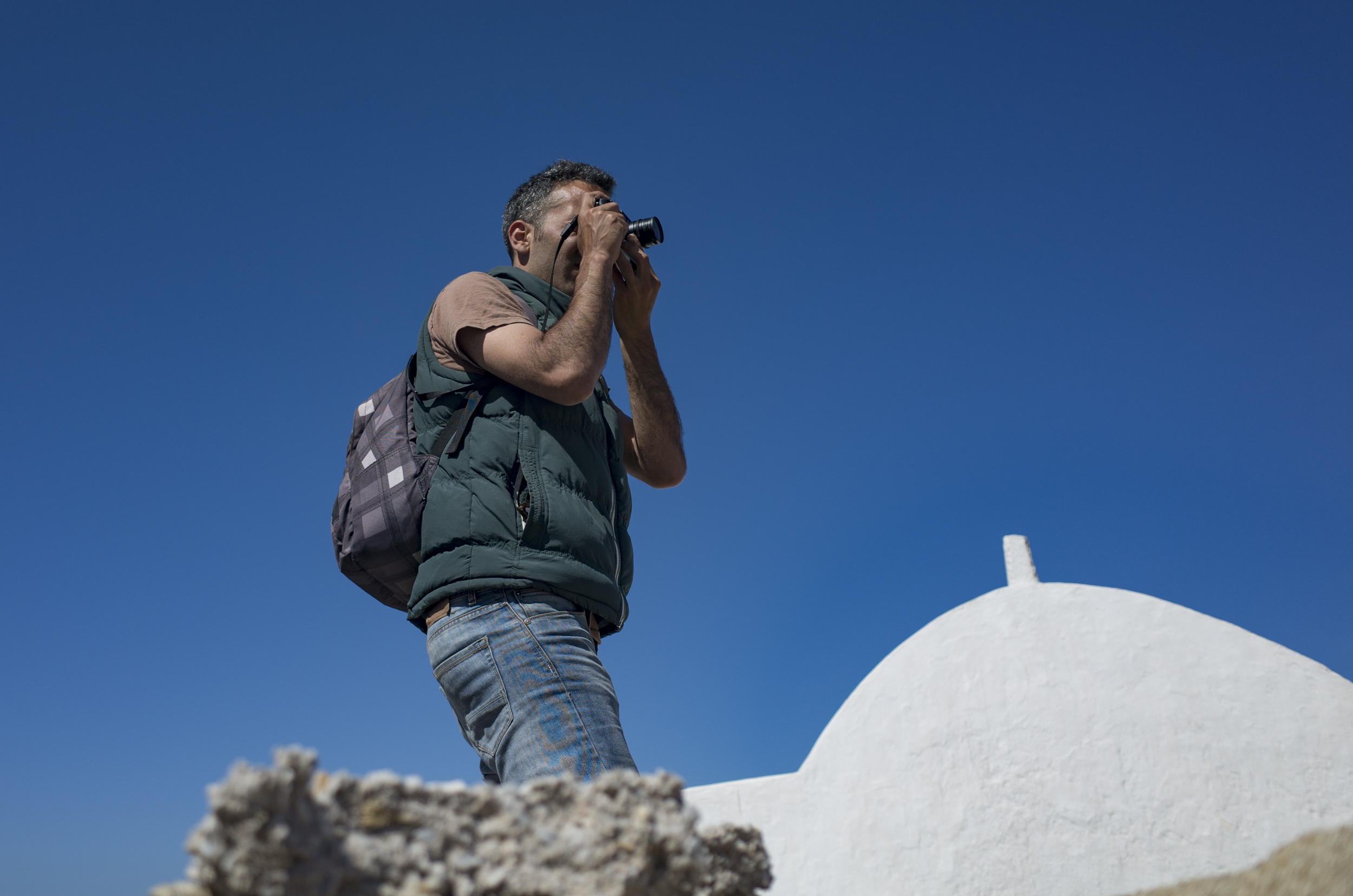A man is standing outdoors, holding a camera to his face, capturing a photograph against a clear blue sky. He wears a green vest and has a checked bag slung over his shoulder.