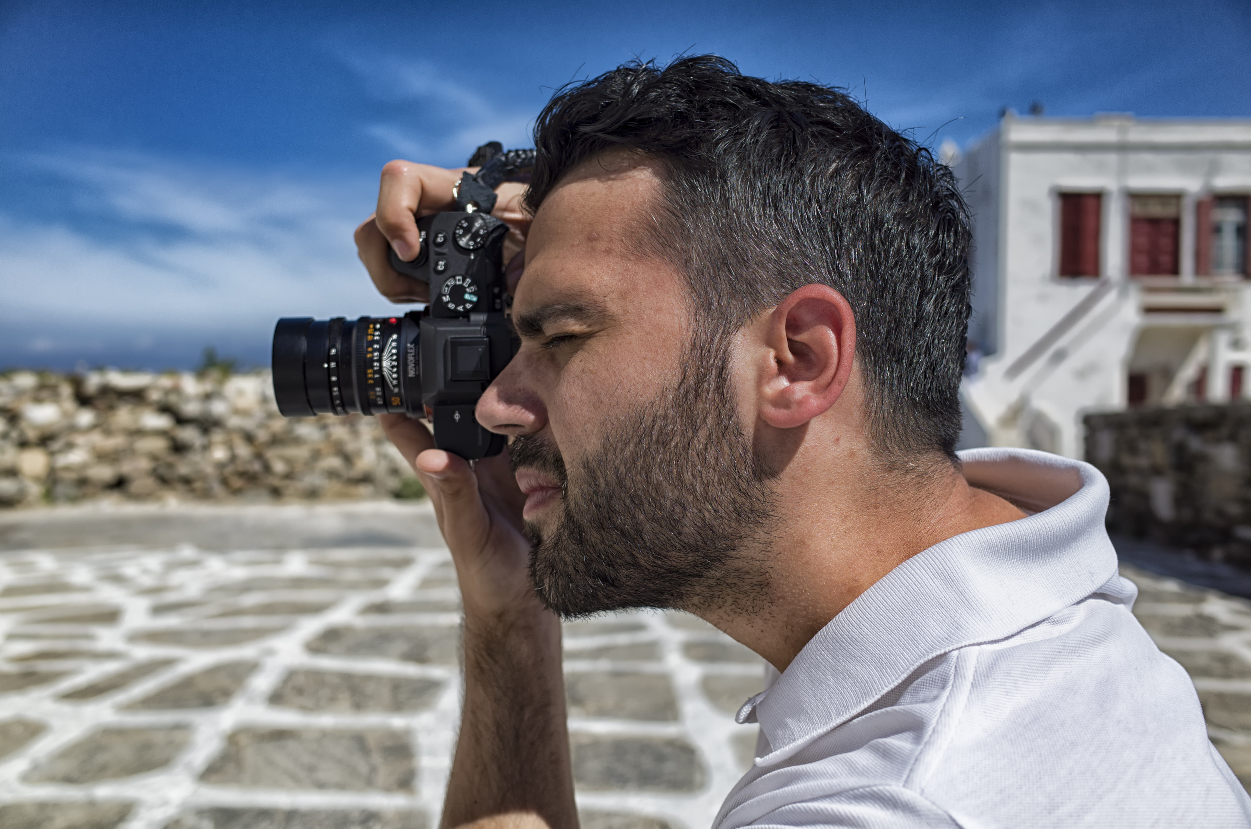 A photographer focusing through a camera, capturing a scene outdoors, with stone pavement and white buildings in the background.
