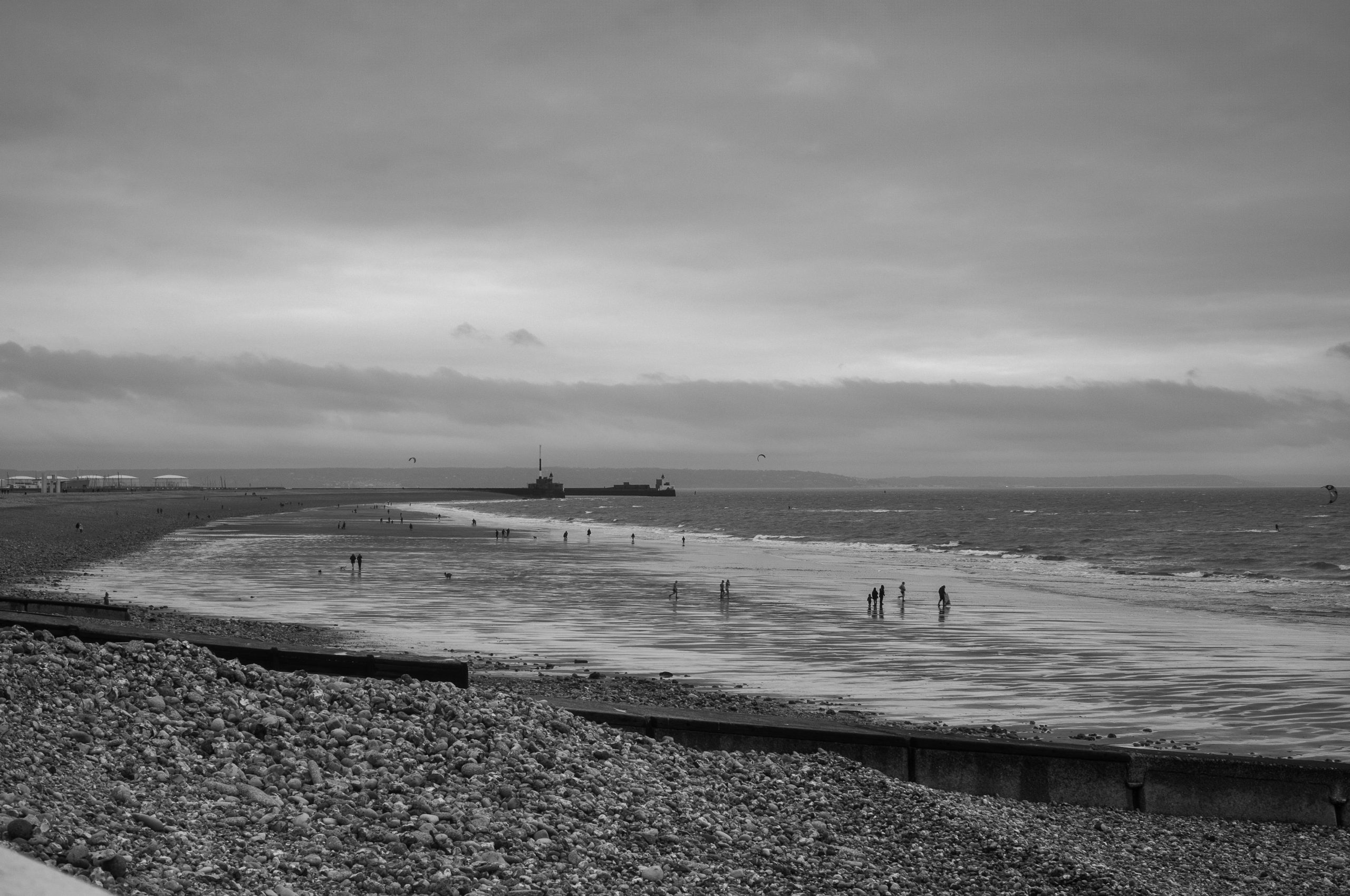 Black and white photograph of a beach scene with people walking along the shore, dotted with water and a distant pier or structure.