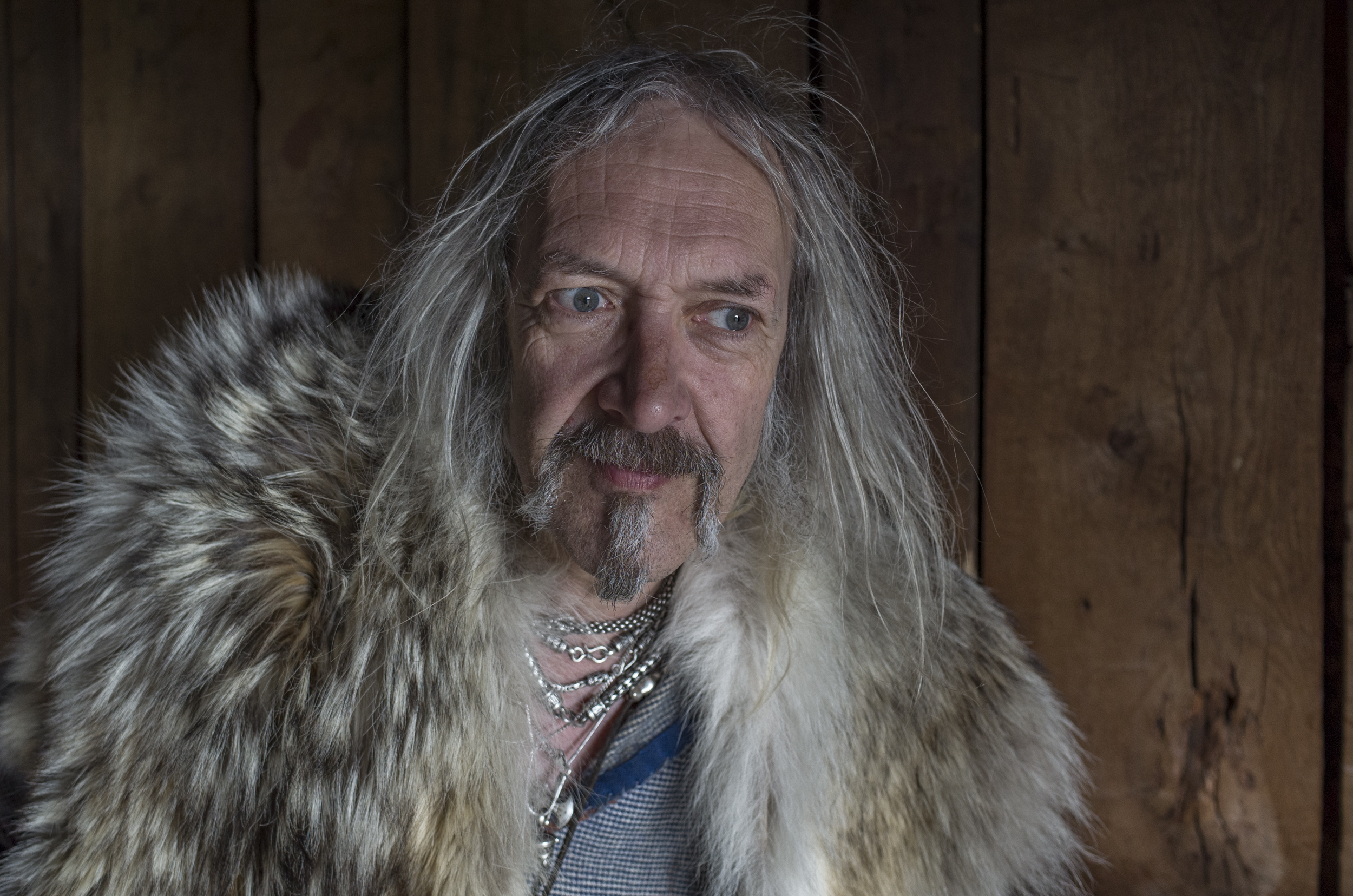 Close-up portrait of a man with long hair, wearing a fur coat, set against a rustic wooden background.