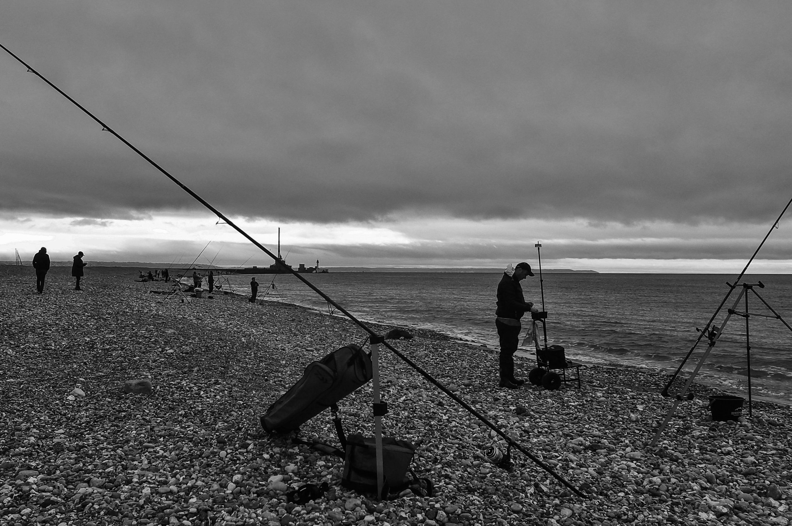 A black and white photo of fishermen along a stony beach, with several individuals casting rods into the water under a cloudy sky.
