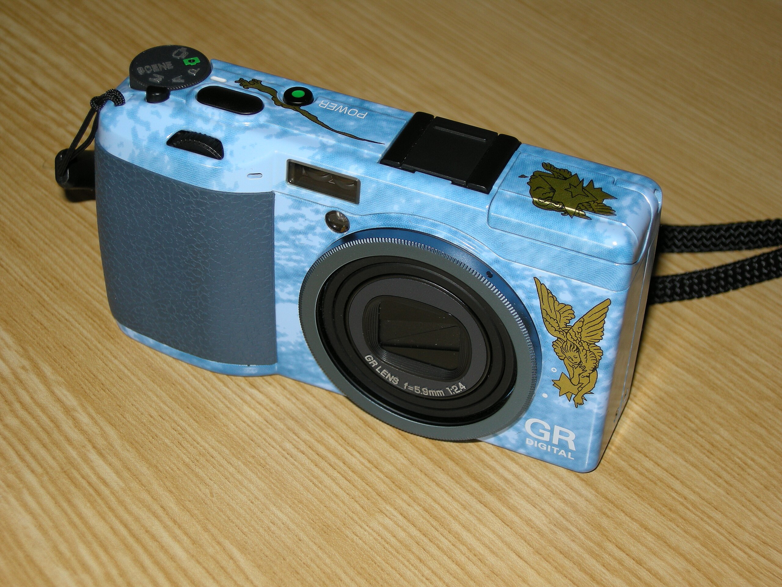 A close-up view of a decorated Ricoh GR Digital camera in a blue design, featuring a textured grip and unique gold graphics, displayed on a wooden surface.