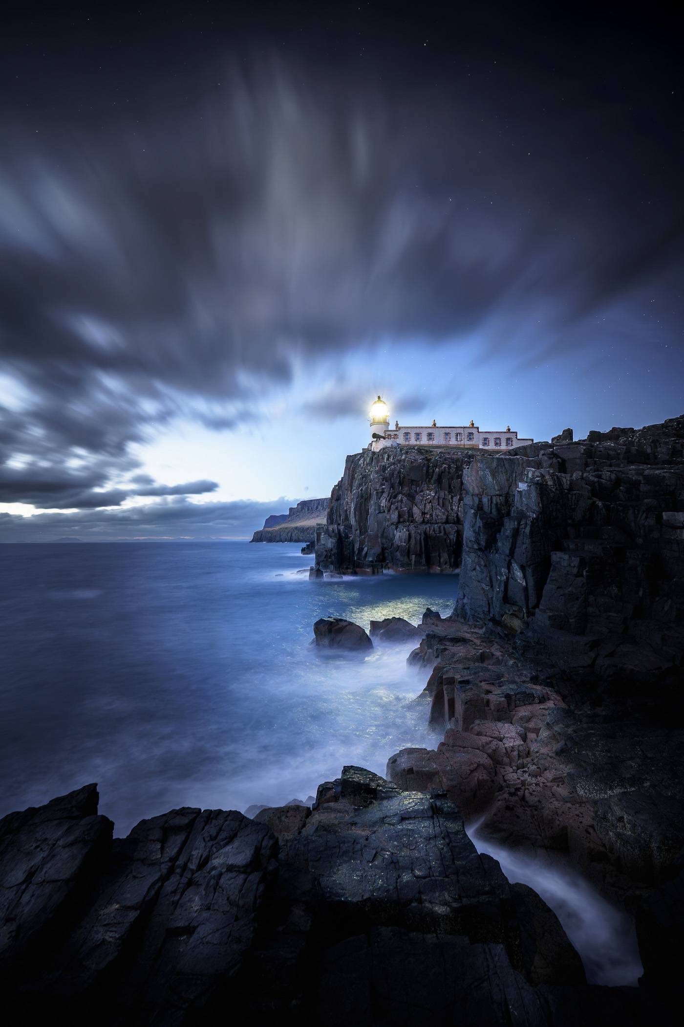 A coastal scene featuring a lighthouse on a cliff, surrounded by rocky shores and dramatic clouds, captured during twilight.