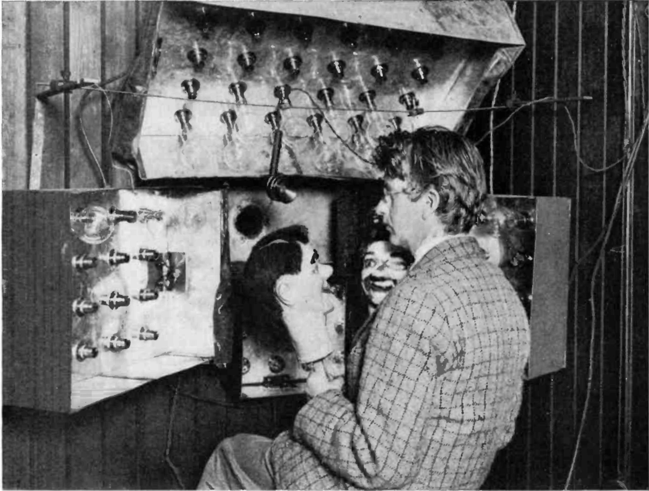 A historic image featuring a man seated in front of an early electromechanical television system, holding a ventriloquist's dummy. The setup includes various vacuum tubes and electrical components in a makeshift studio environment.