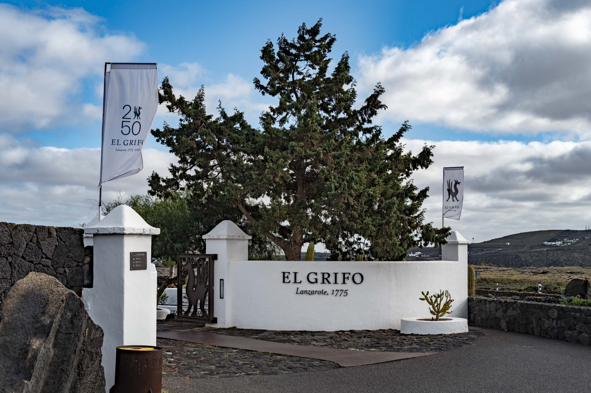 Entrance to El Grifo winery in Lanzarote, featuring a white wall with signage and a flag, surrounded by volcanic rock and greenery.
