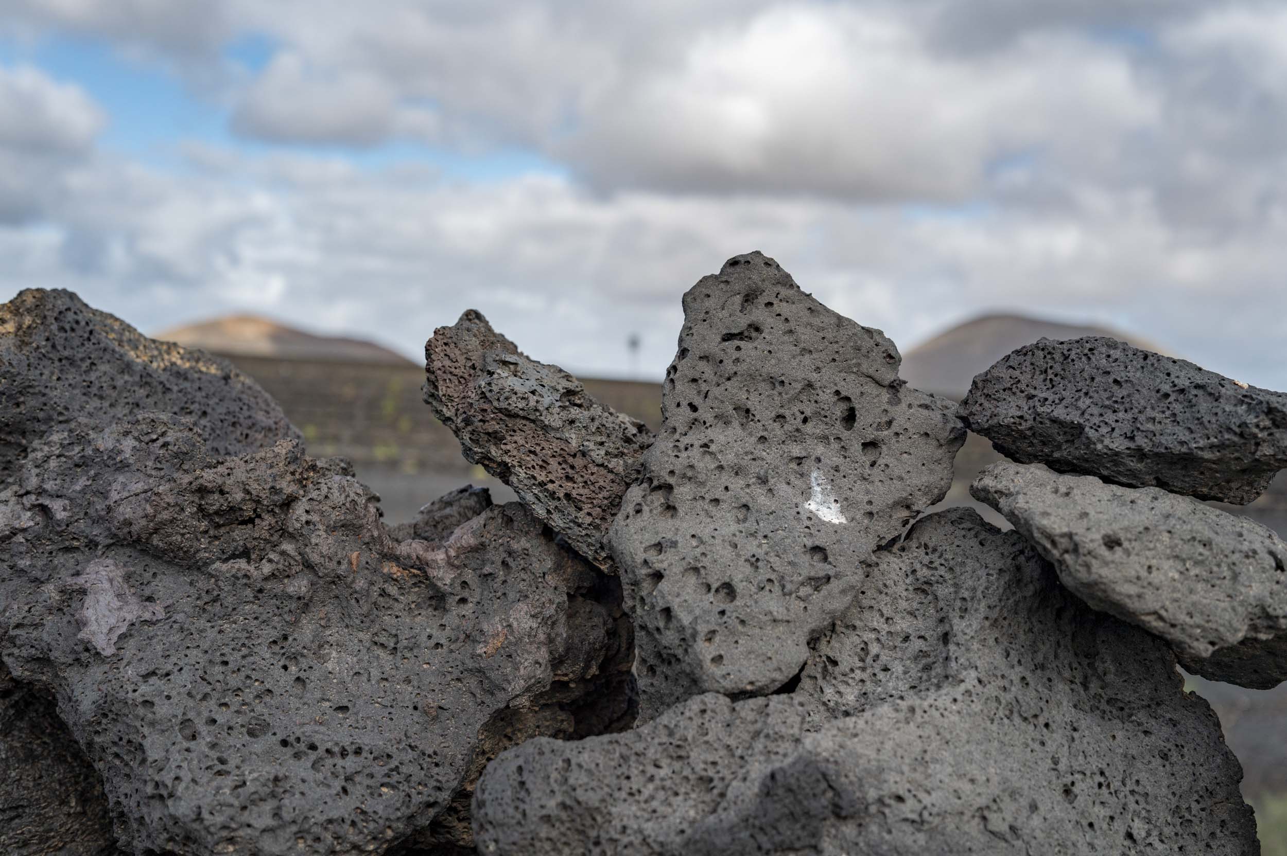 Close-up of volcanic rocks with a blurred background of Lanzarote's landscape under cloudy skies.