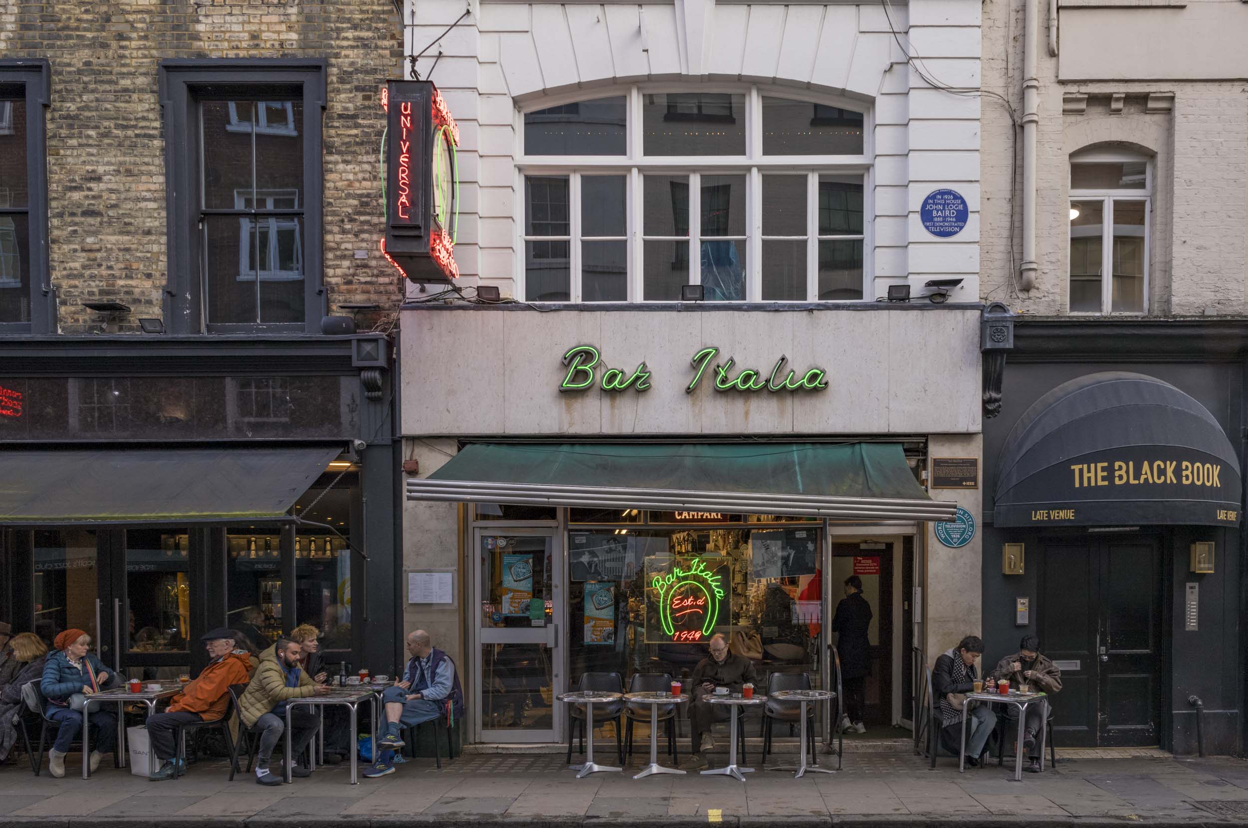 Exterior view of Bar Italia, a historic Italian coffee bar in London, featuring neon signage and patrons seated outside.