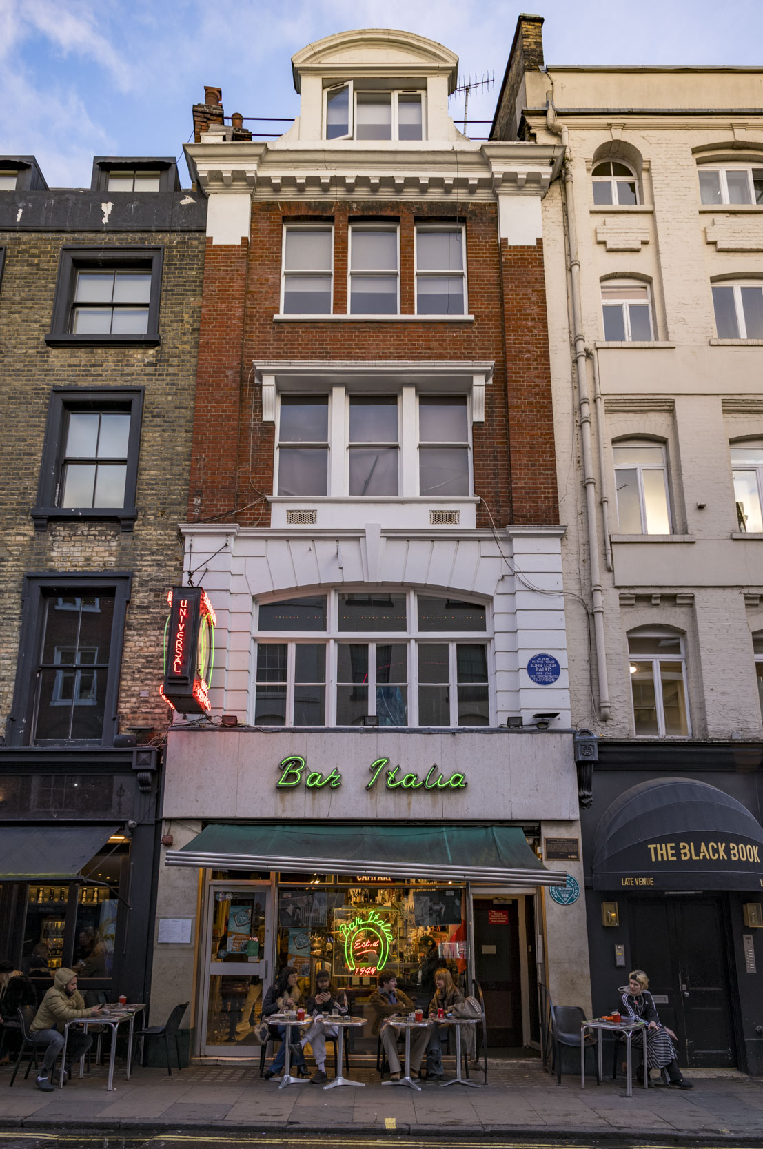 Exterior view of Bar Italia, a café with a neon sign, featuring outdoor seating and people enjoying drinks on a street in London.