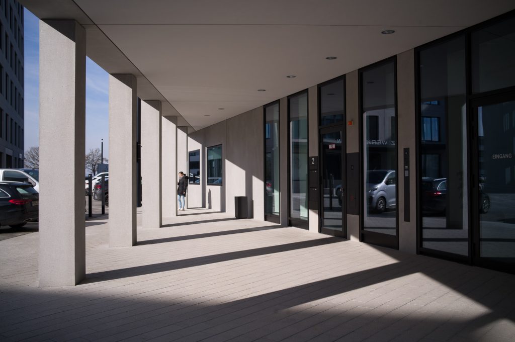 A modern architectural corridor featuring tall concrete columns, large glass windows, and a person standing near the building entrance. Bright sunlight creates distinct shadows on the ground.