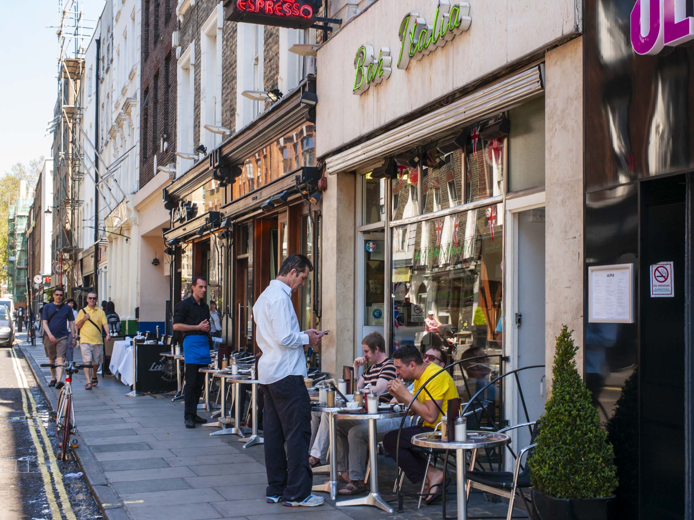 Exterior view of Bar Italia in Soho, London, with patrons sitting at tables outside, a waiter attending to customers, and a vibrant street scene.