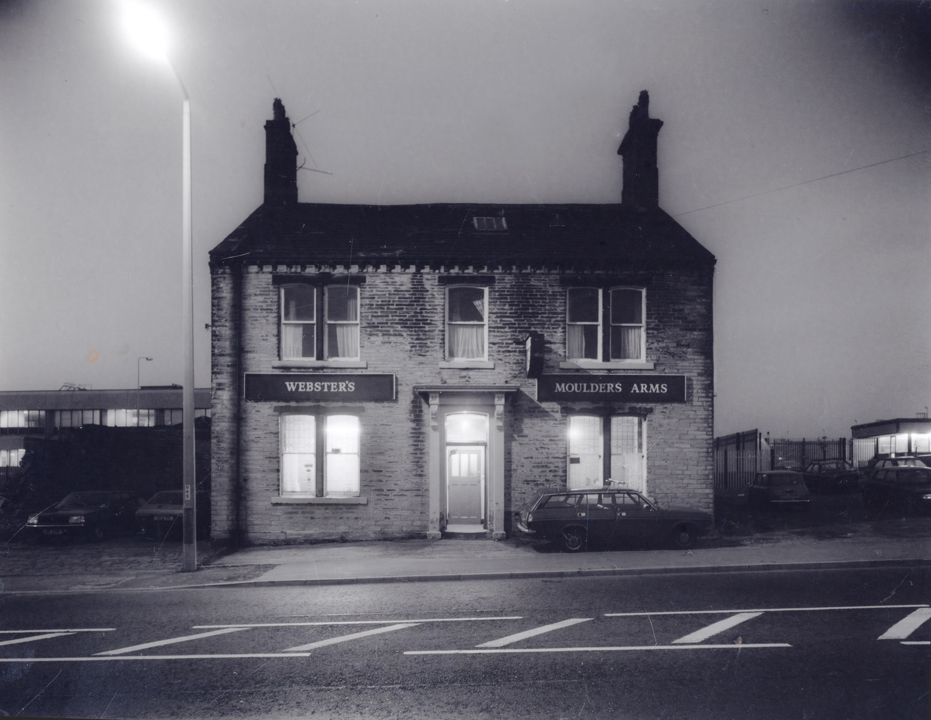 Black and white photograph of the Moulders Arms pub with a sign that reads 'Webster's' in front, taken at dusk with street lighting illuminating the building, parked cars visible on the street.