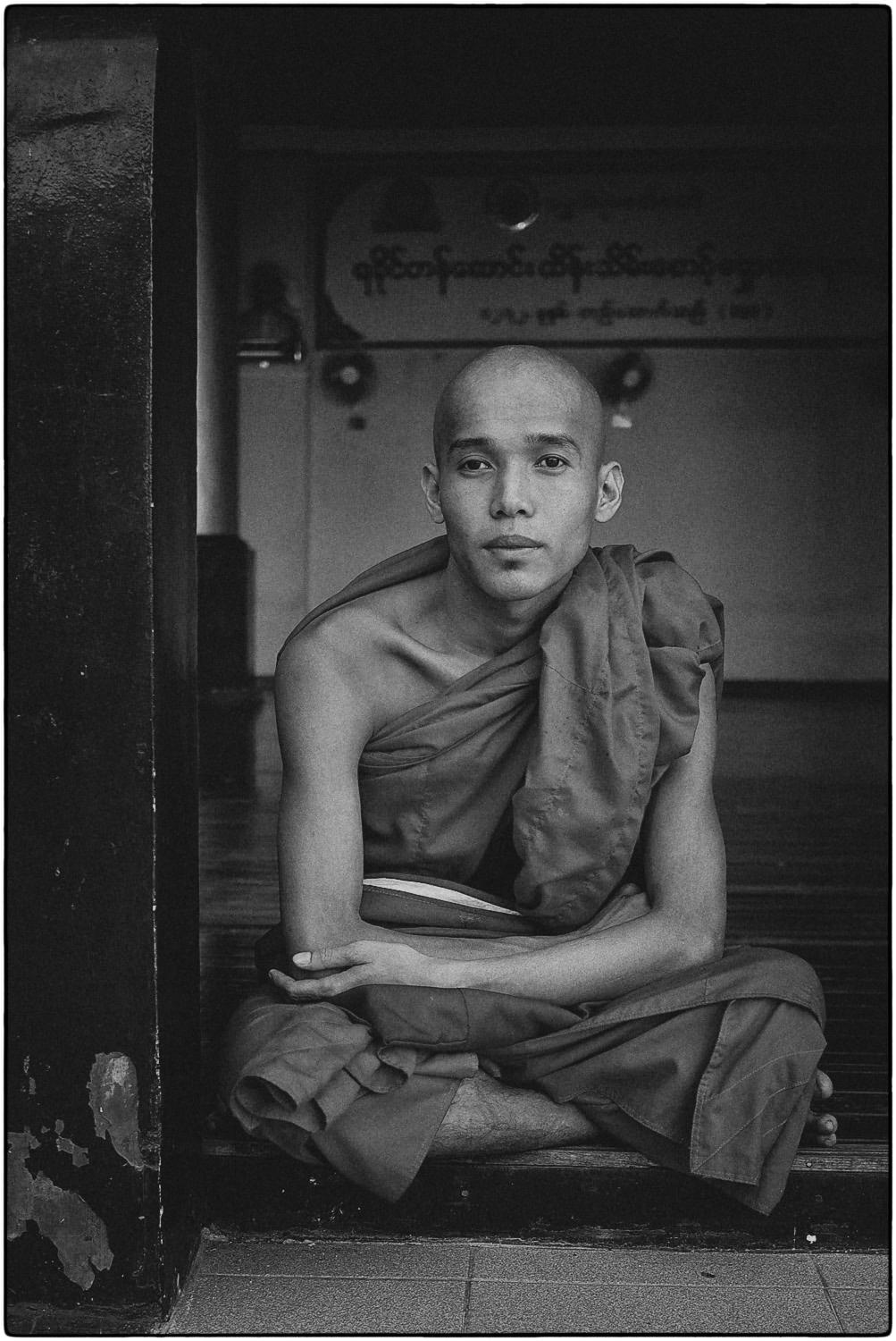 A young monk in traditional robes sitting cross-legged in a temple setting, with a contemplative expression.