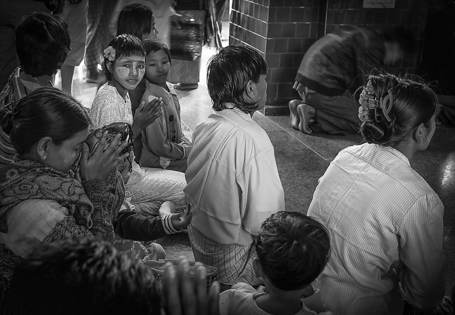 A group of people, including children, sitting together in a prayer-like posture, some with hands clasped. The image is in black and white, with individuals wearing traditional clothing, set in an indoor environment.