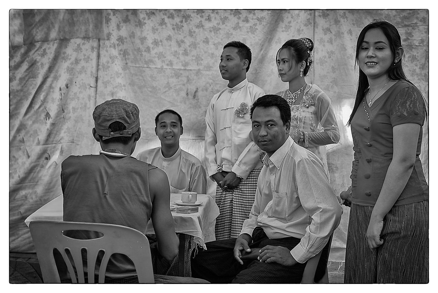 A group of five people posing for a photo in front of a light-coloured backdrop. Two men are seated at a table, while three others stand behind them, all smiling. The scene is in black and white.