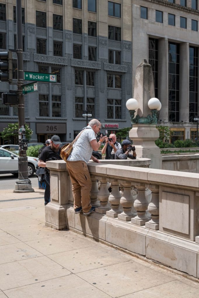A group of photographers capturing images near a decorative stone railing in an urban setting, with buildings and street signs visible in the background.