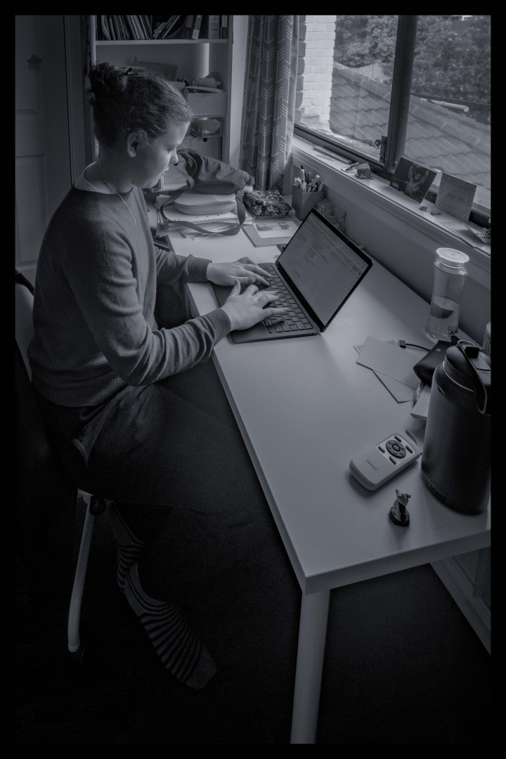 A person sitting at a desk, typing on a laptop with a window in the background. The image is in black and white, showing a tidy workspace with books, stationery, and a water bottle on the desk.