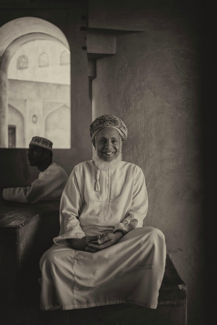 A smiling older man in traditional dress sits cross-legged against a wall in a historic building, with another figure partially visible in the background.