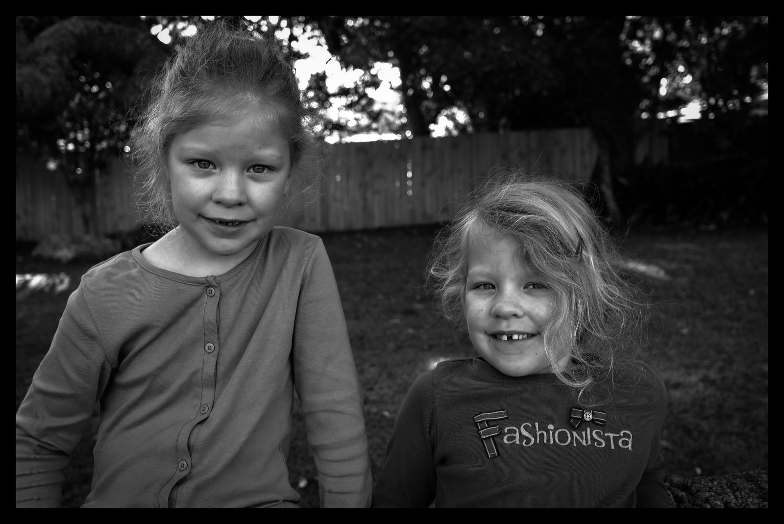 Two young girls smiling at the camera, one wearing a blue top and the other a top with the word 'Fashionista'. The background shows a garden with a wooden fence.