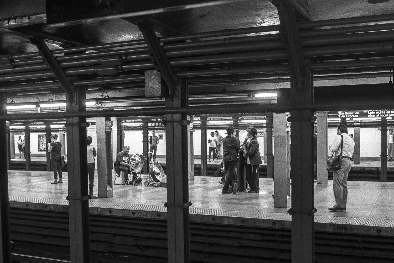 A black and white image of a subway station platform, featuring several people waiting for a train, with one person playing a drum in the foreground.