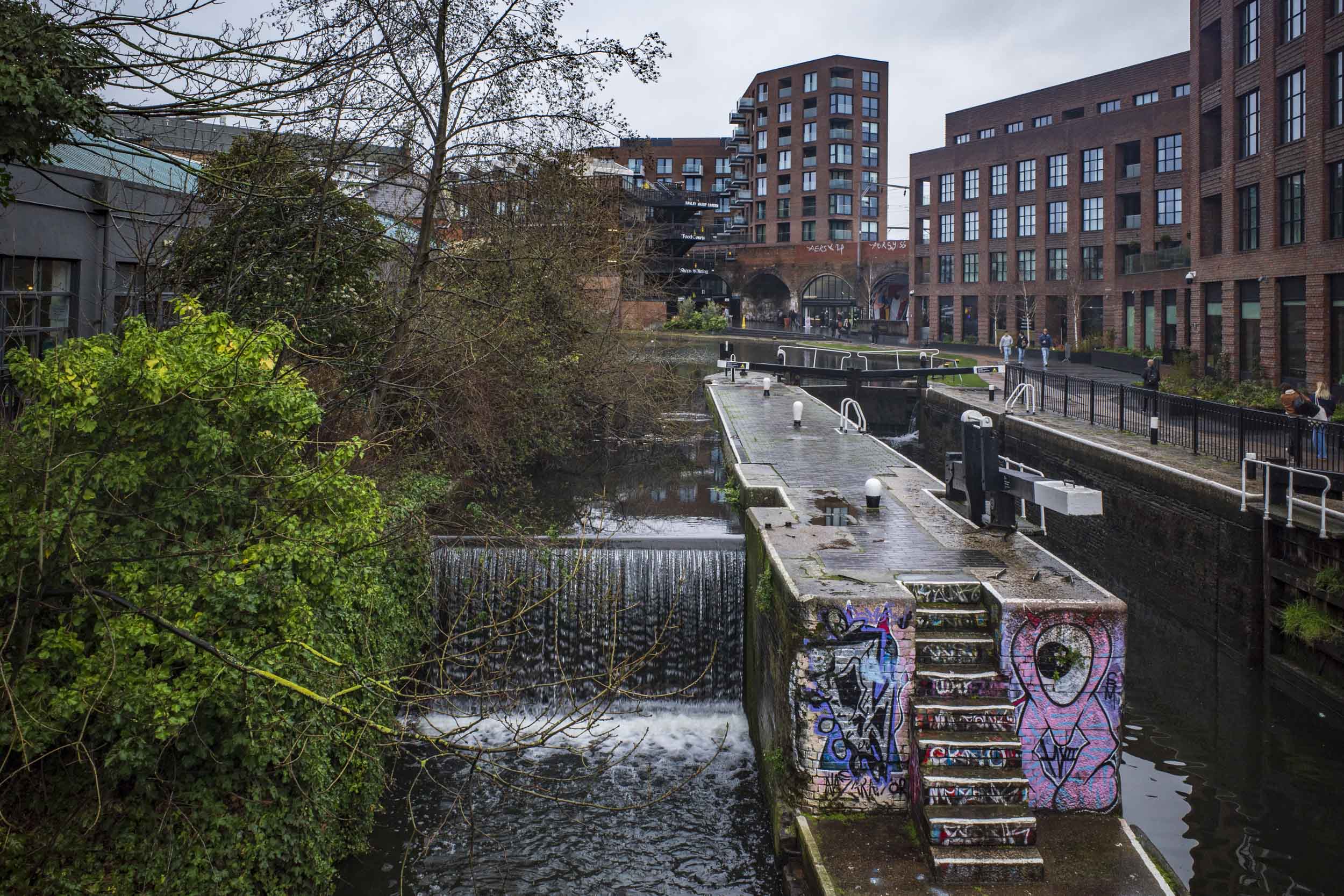 A canal scene featuring a small waterfall, steps adorned with graffiti, and modern brick buildings in the background. Overcast skies indicate a cloudy day.