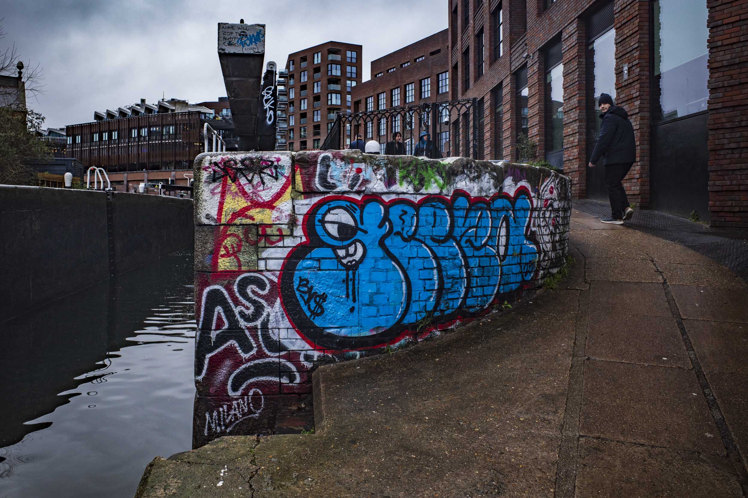 A canal bank adorned with vibrant graffiti, featuring large blue lettering, alongside a paved walkway. In the background, modern buildings are visible under a cloudy sky.