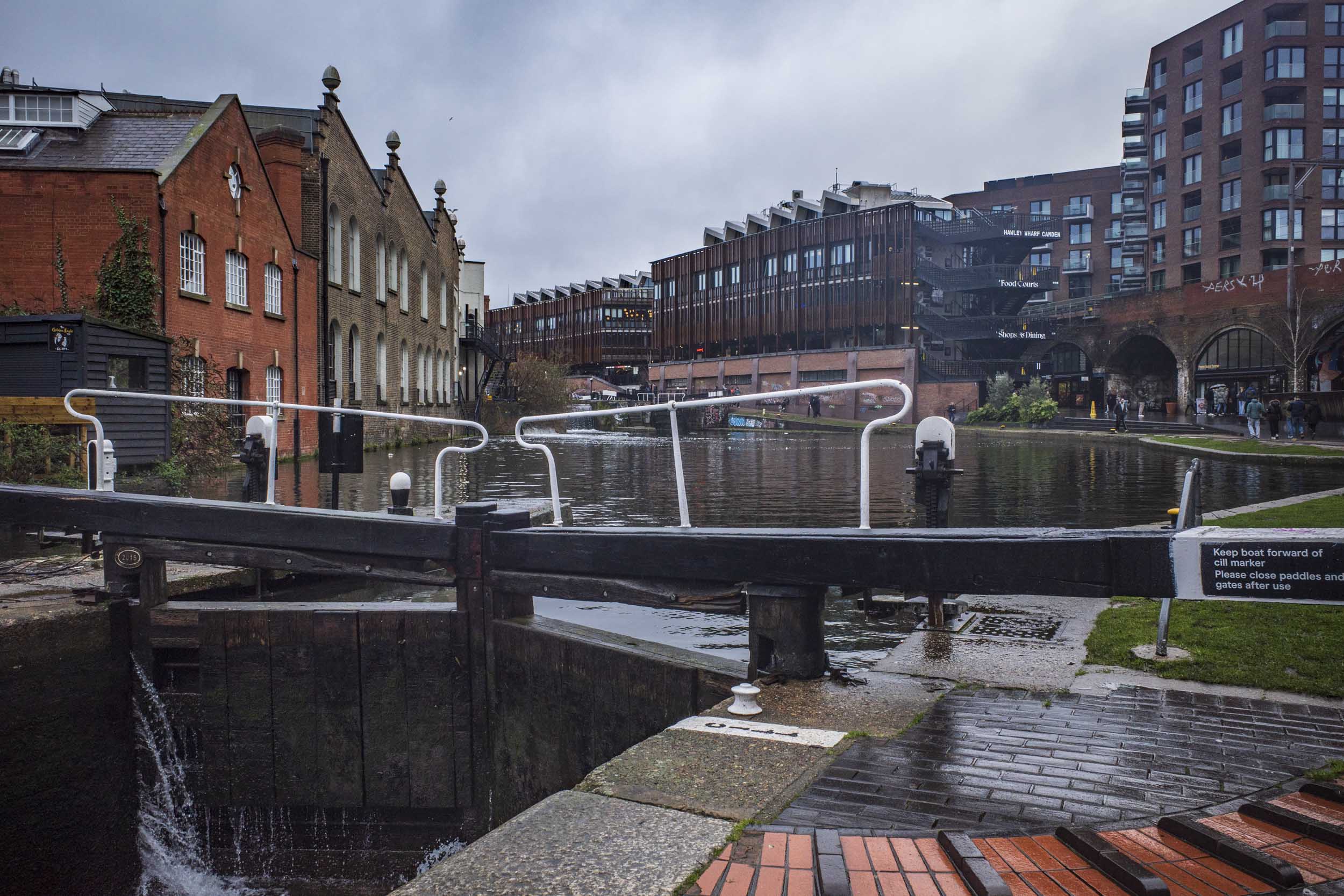 View of a canal with a lock gate in the foreground, surrounded by brick and modern buildings, under a cloudy sky.
