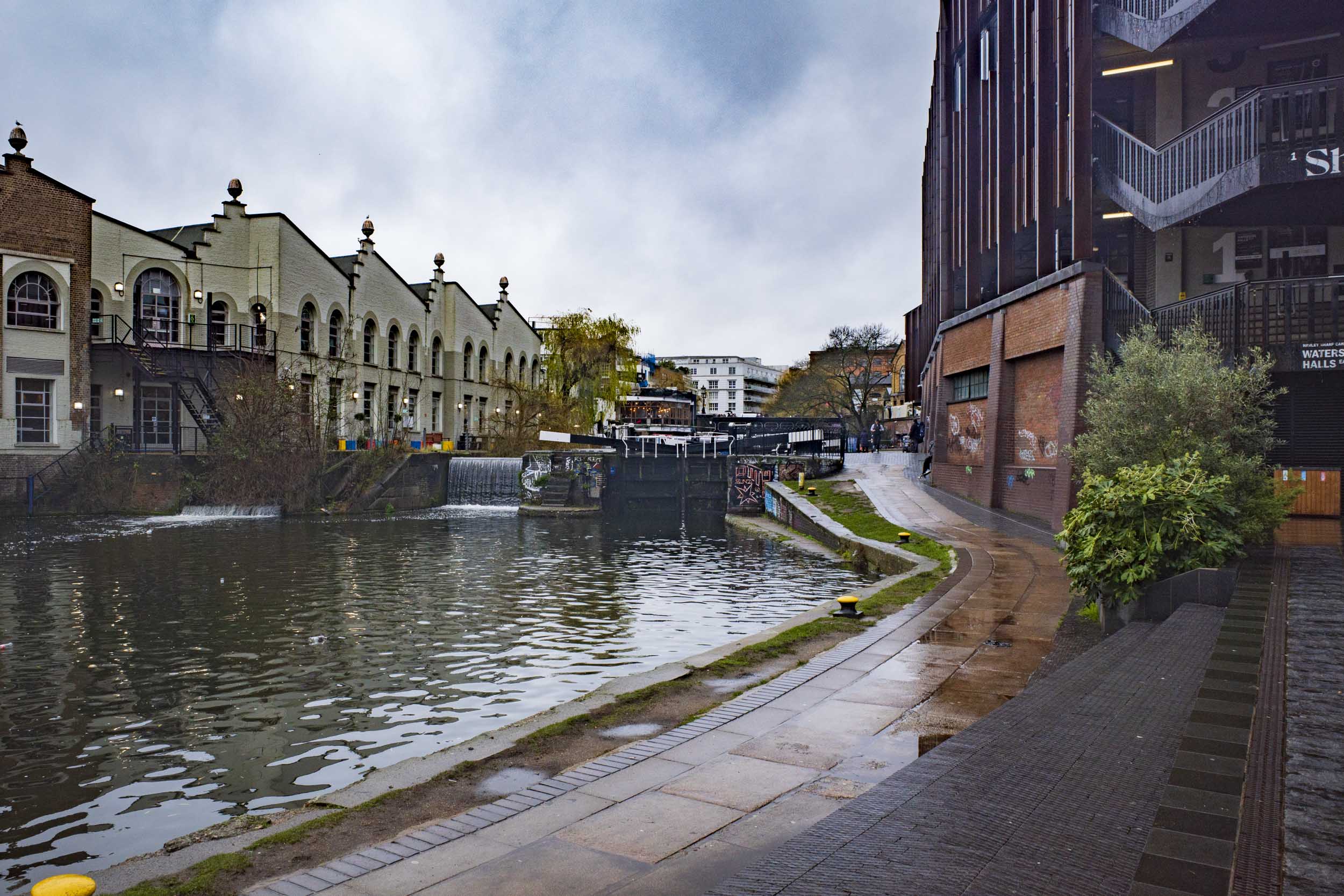View of a canal with a waterfall, flanked by historical buildings and modern architecture on a cloudy day.