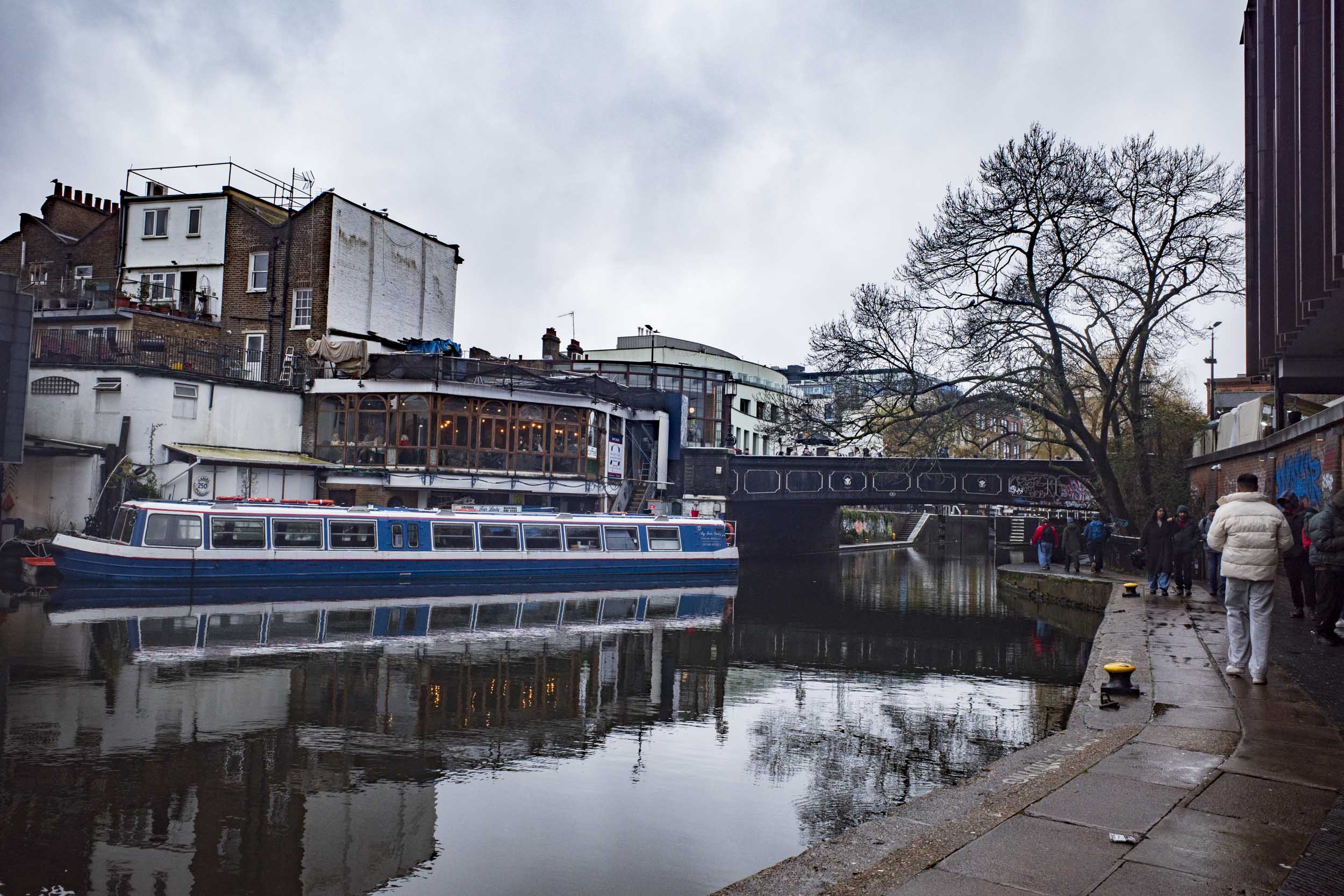 A tranquil scene along a canal with moored boats, reflecting the surrounding buildings. A bridge crosses the water, with people walking along the bank under a cloudy sky.