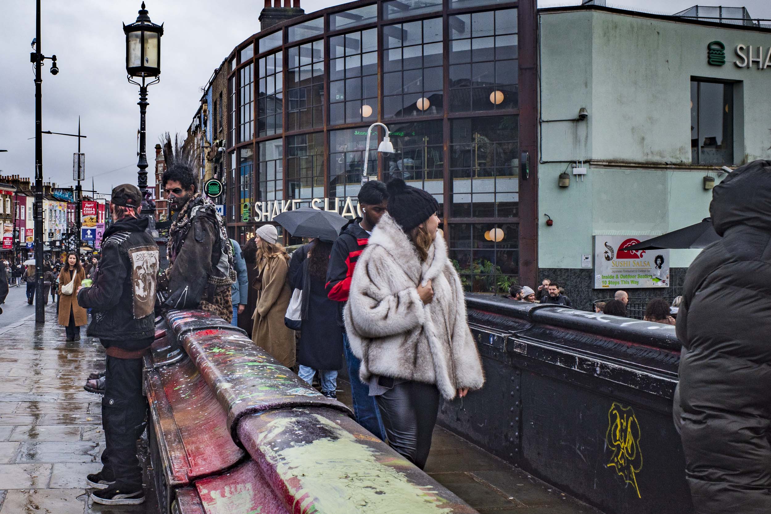A bustling street scene featuring pedestrians in stylish outfits, including a woman in a furry coat, near shops and eateries in an urban setting.