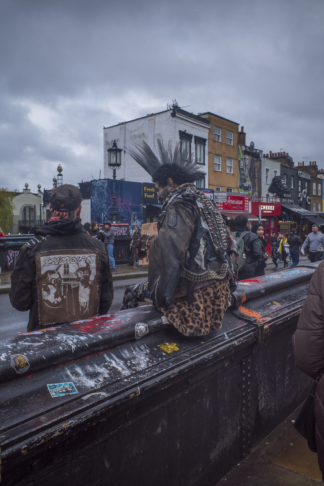 Two individuals with distinctive punk fashion sitting on a railing in a busy urban setting. One person has spiky hair and a leather jacket, while the other has a cap and a graphic back patch. The background features buildings and street life under a cloudy sky.