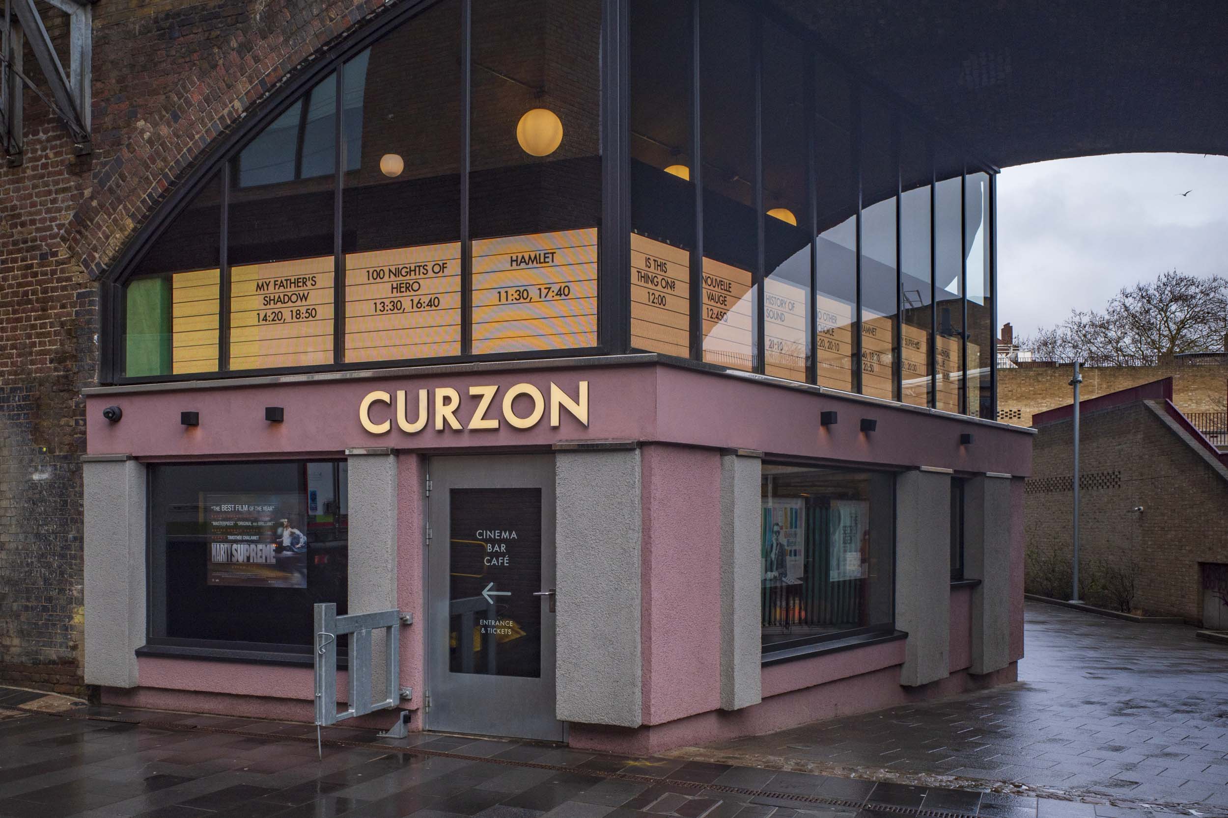 Exterior view of the Curzon cinema, featuring large glass windows and a sign displaying film titles and showtimes. The building is located under an archway, with a modern design and nearby pavement.