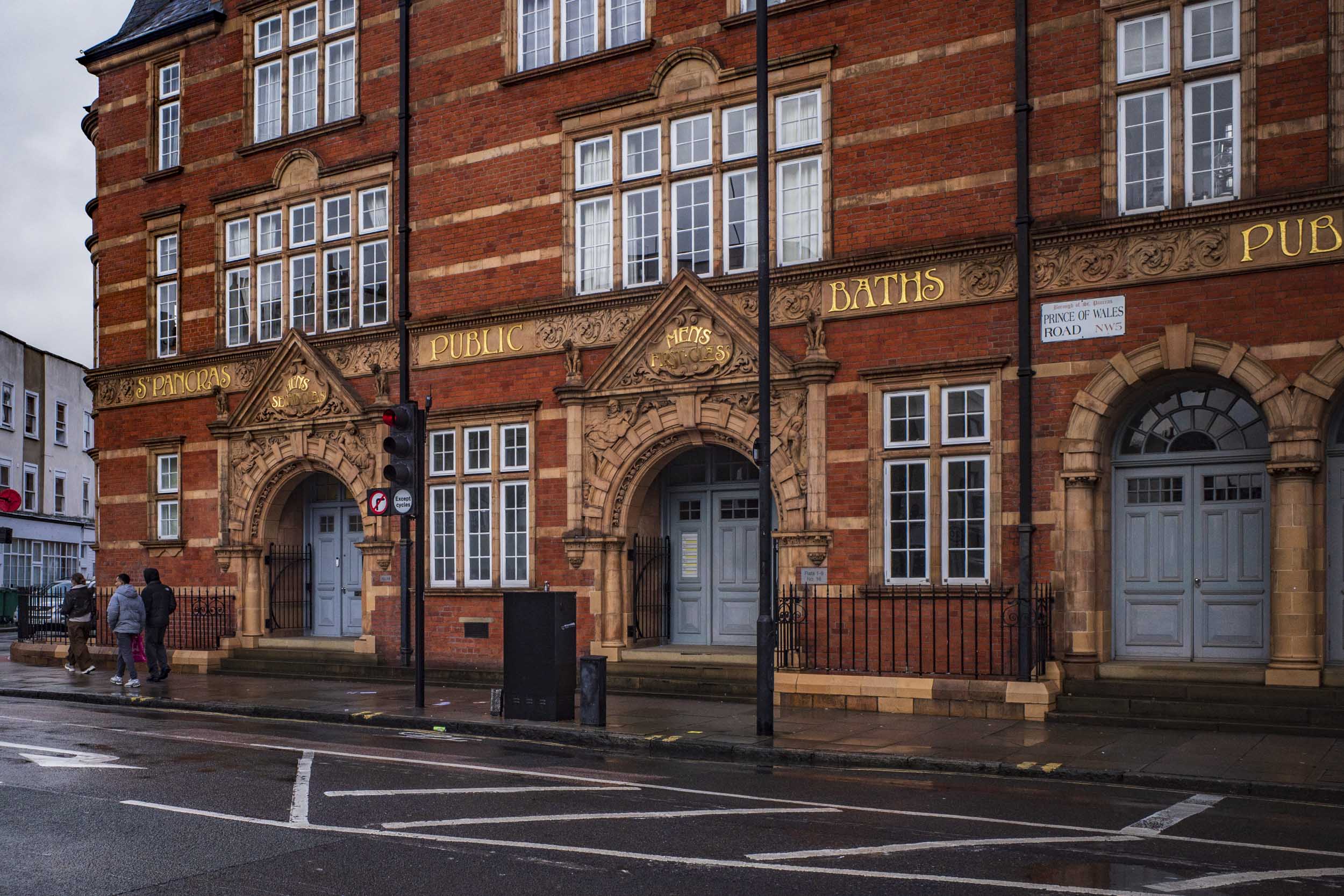 Historic building featuring ornamental architecture, labelled 'St Pancras Public Baths', with pedestrians walking on the street and traffic signals visible.