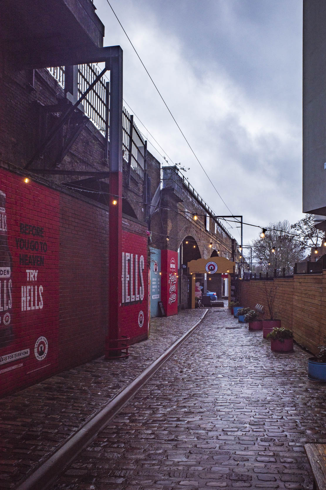 A cobbled alleyway lined with brick walls featuring vibrant advertisements, under a cloudy sky. Potted plants decorate the space, creating a quaint atmosphere.