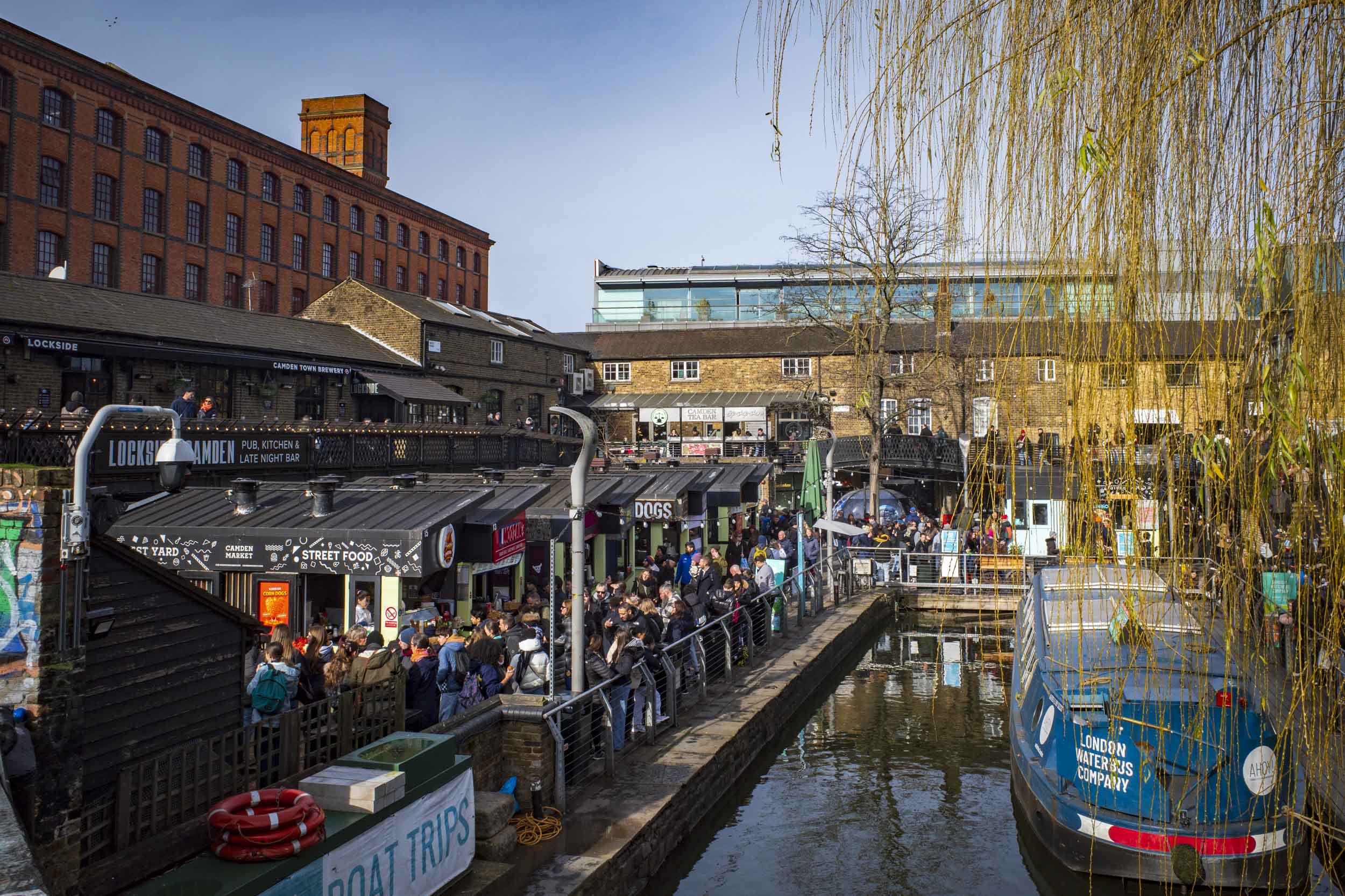 A bustling scene at Camden Market, with many people enjoying the shops and eateries along the canal. A boat is docked on the water, and historic buildings provide a backdrop. The area is lively with a mix of street food stalls and outdoor seating.