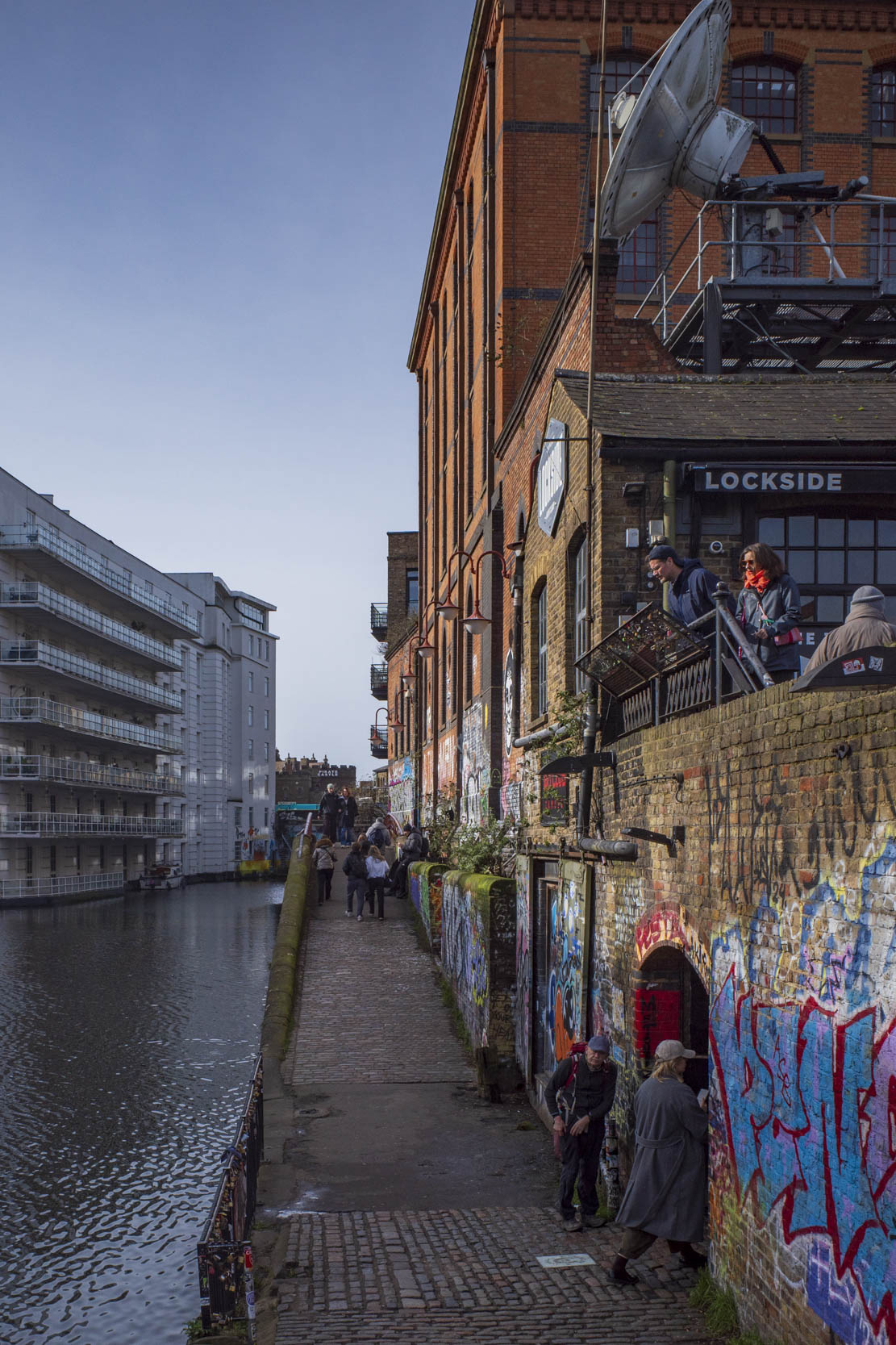 A scenic view of a cobbled walkway beside a canal, featuring graffiti-covered brick walls and buildings. People are walking along the path, while others are seated on a balcony above, enjoying the surroundings on a clear day.