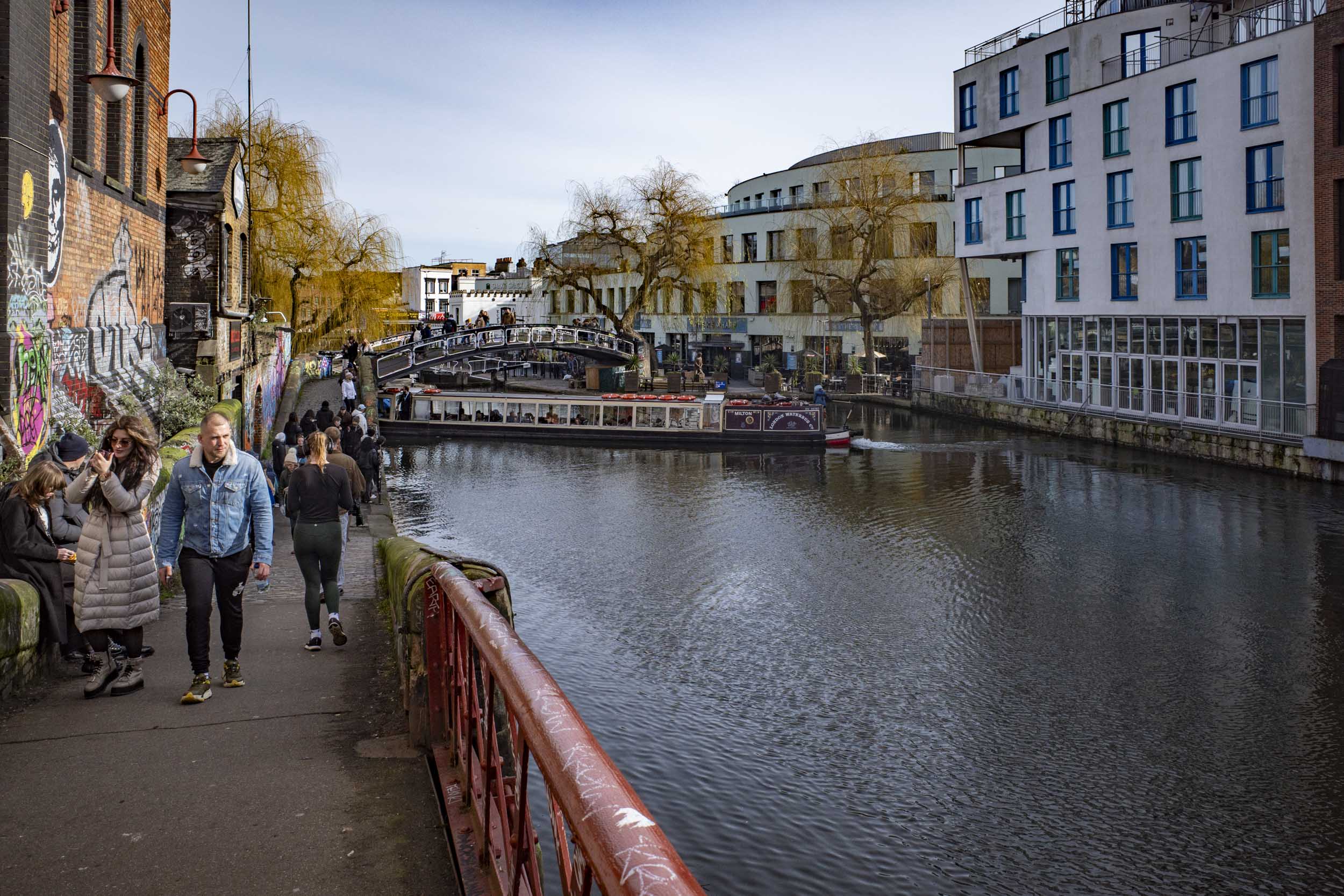 A bustling canal scene with people walking along a path beside the water, narrowboats moored along the canal, and modern buildings alongside, featuring a mixture of brick and glass architecture.