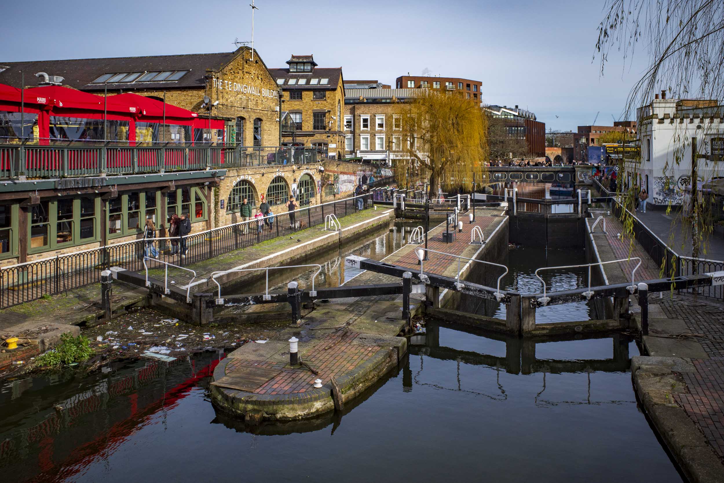 A scenic view of a canal with locks, surrounded by historic buildings and a restaurant with a red awning. Pedestrians walk along the canal path.