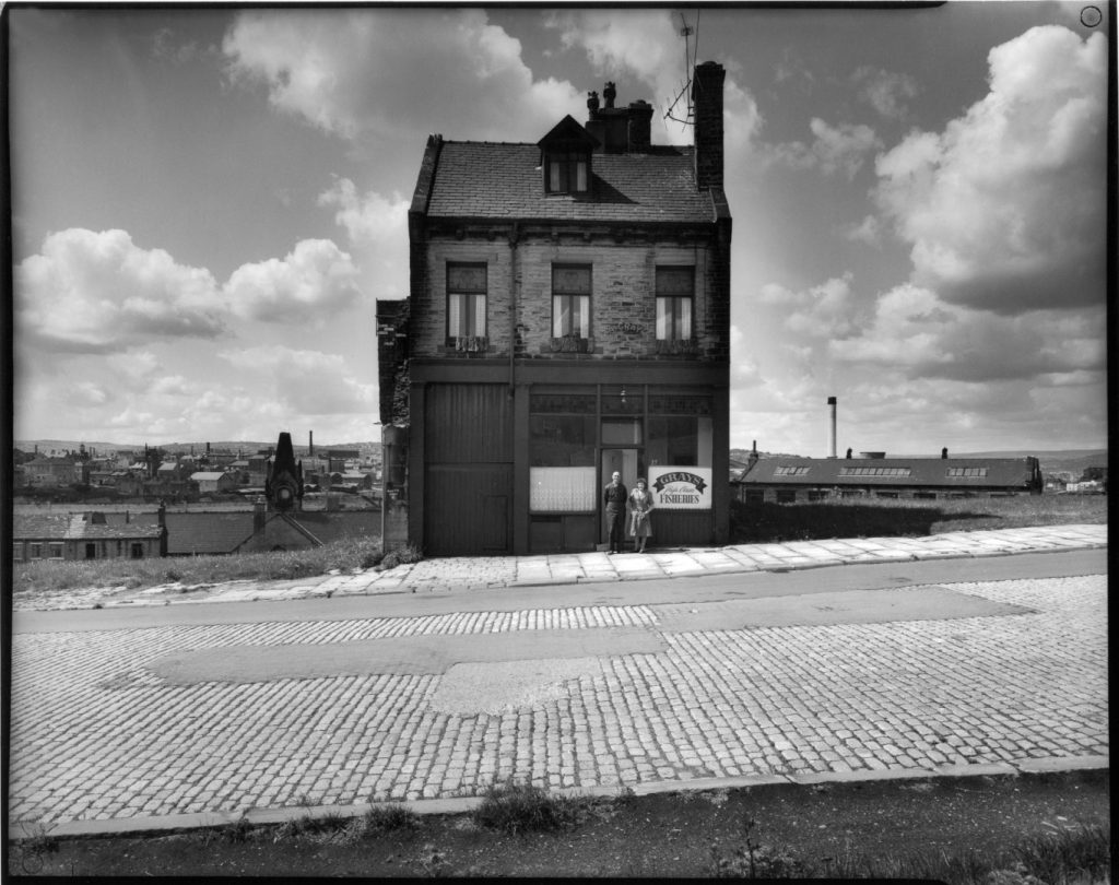 Black and white photograph of a two-storey building with a sign reading 'Fisheries', featuring two people standing at the entrance. The surrounding area includes cobblestone pavement and a distant industrial skyline under a partly cloudy sky.
