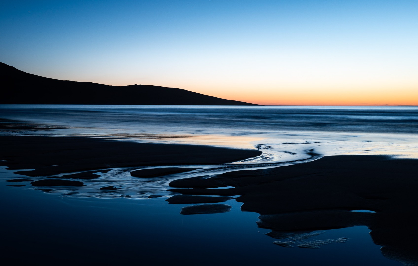 A serene beach scene at dusk, featuring gently flowing water and a smooth shoreline, with a silhouetted mountain in the background and a gradient sky transitioning from blue to orange.